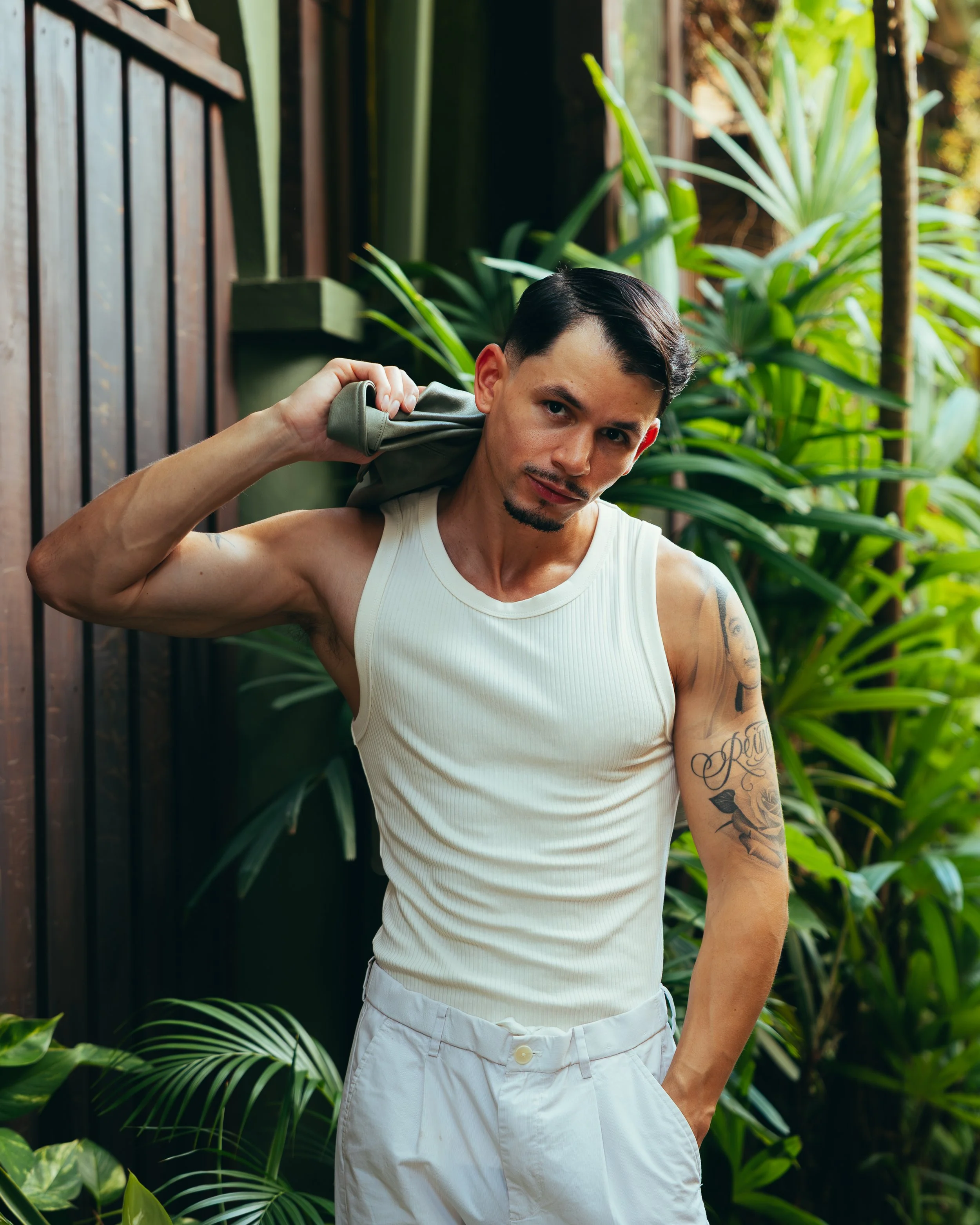A man carrying a gray backpack over his shoulder stands in front of green tropical plants, wearing a white sleeveless shirt and white pants. He has tattoos on his left arm and dark, styled hair.