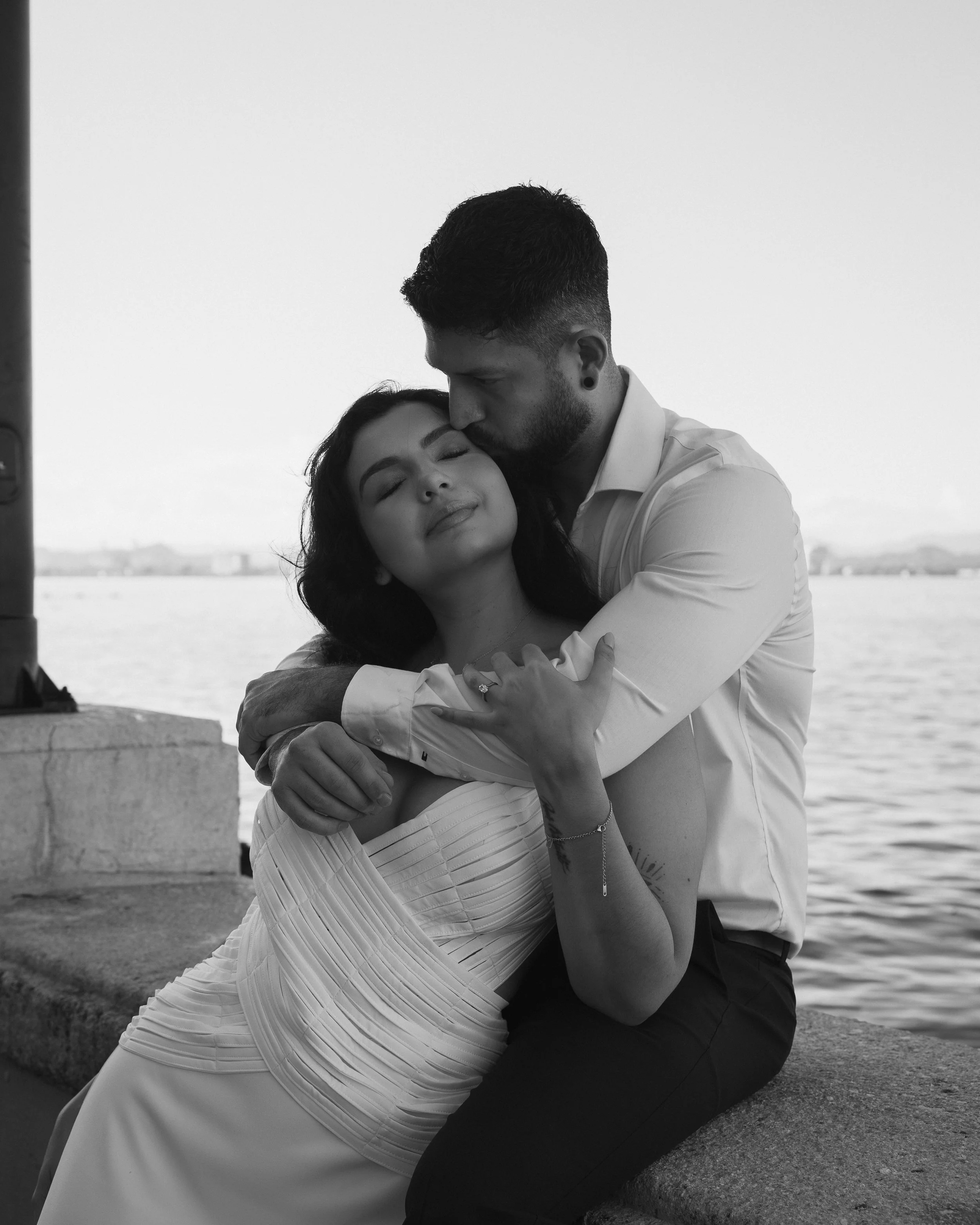 A black and white photo of a couple embracing by a body of water, with the man giving a gentle kiss to the woman's forehead and the woman smiling with her eyes closed.