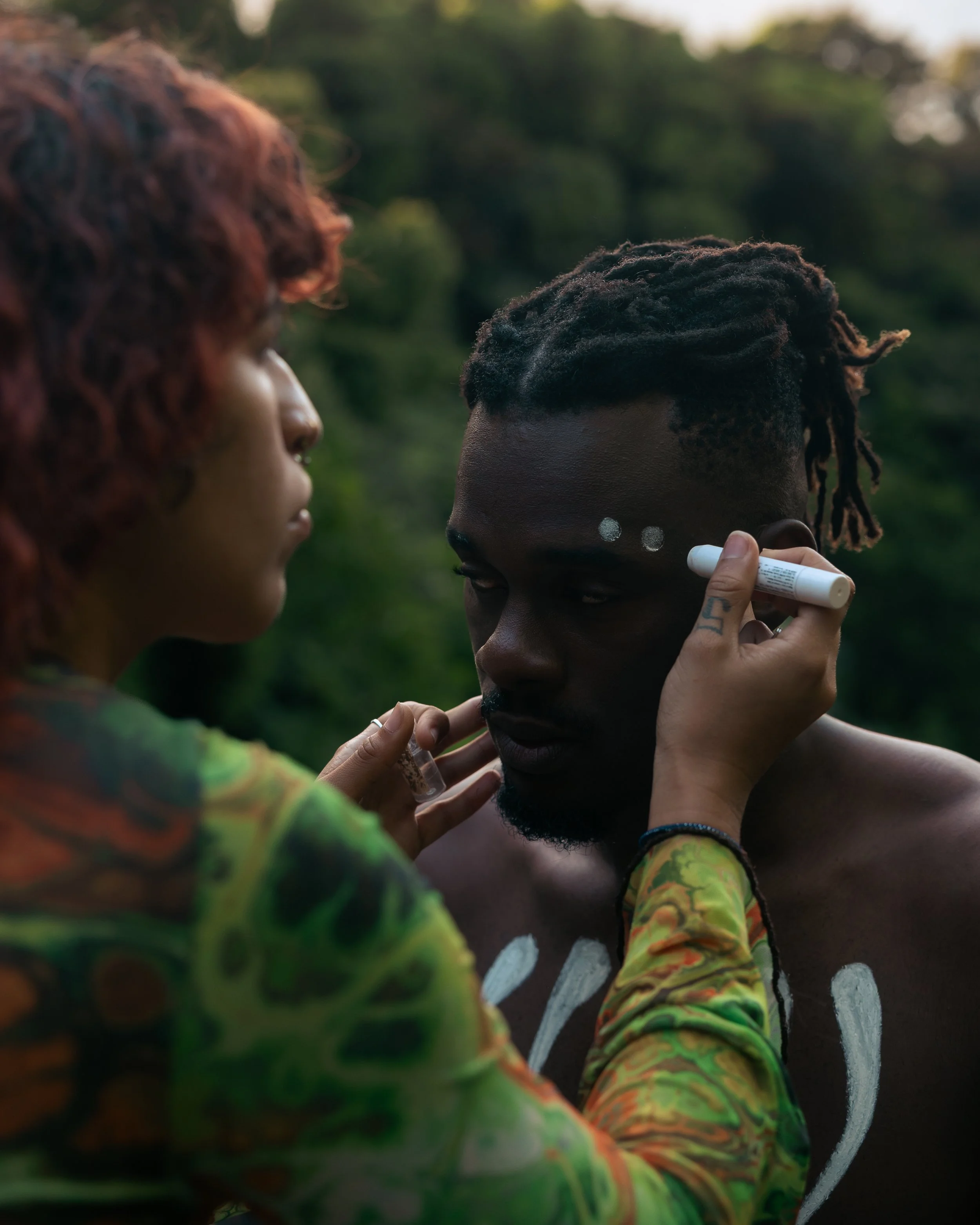 A woman with reddish hair paints white dots on a shirtless man with dark skin and dreadlocks outdoors during daytime, with a blurred green background.