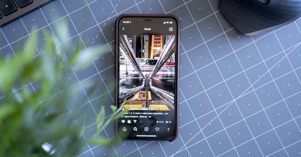 Smartphone displaying Instagram Reels on a grid-patterned desk next to a leafy plant and a mouse.