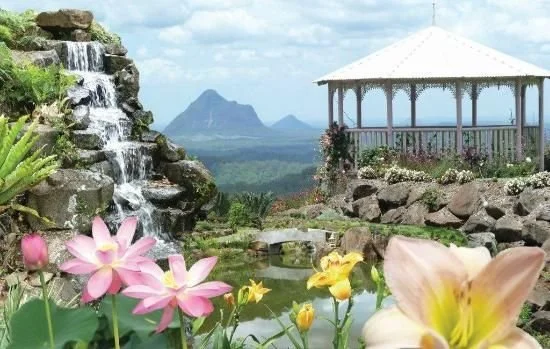 Scenic garden with a waterfall, colorful flowers, a pond, a small bridge, and a gazebo against a mountain backdrop.