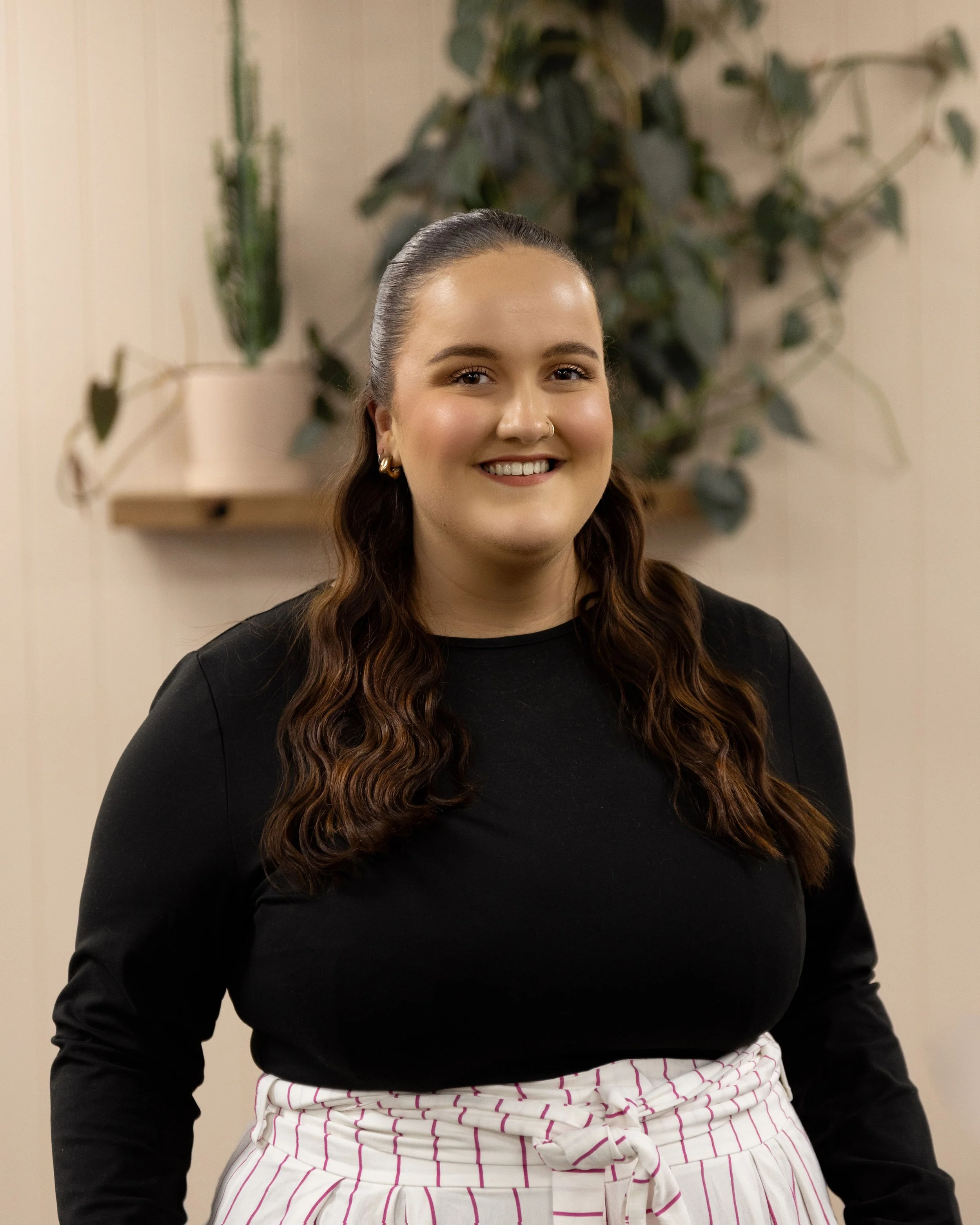 A woman with long, wavy brown hair, smiling, wearing a black top and white and pink striped pants, standing in front of a beige wall with a plant in the background.