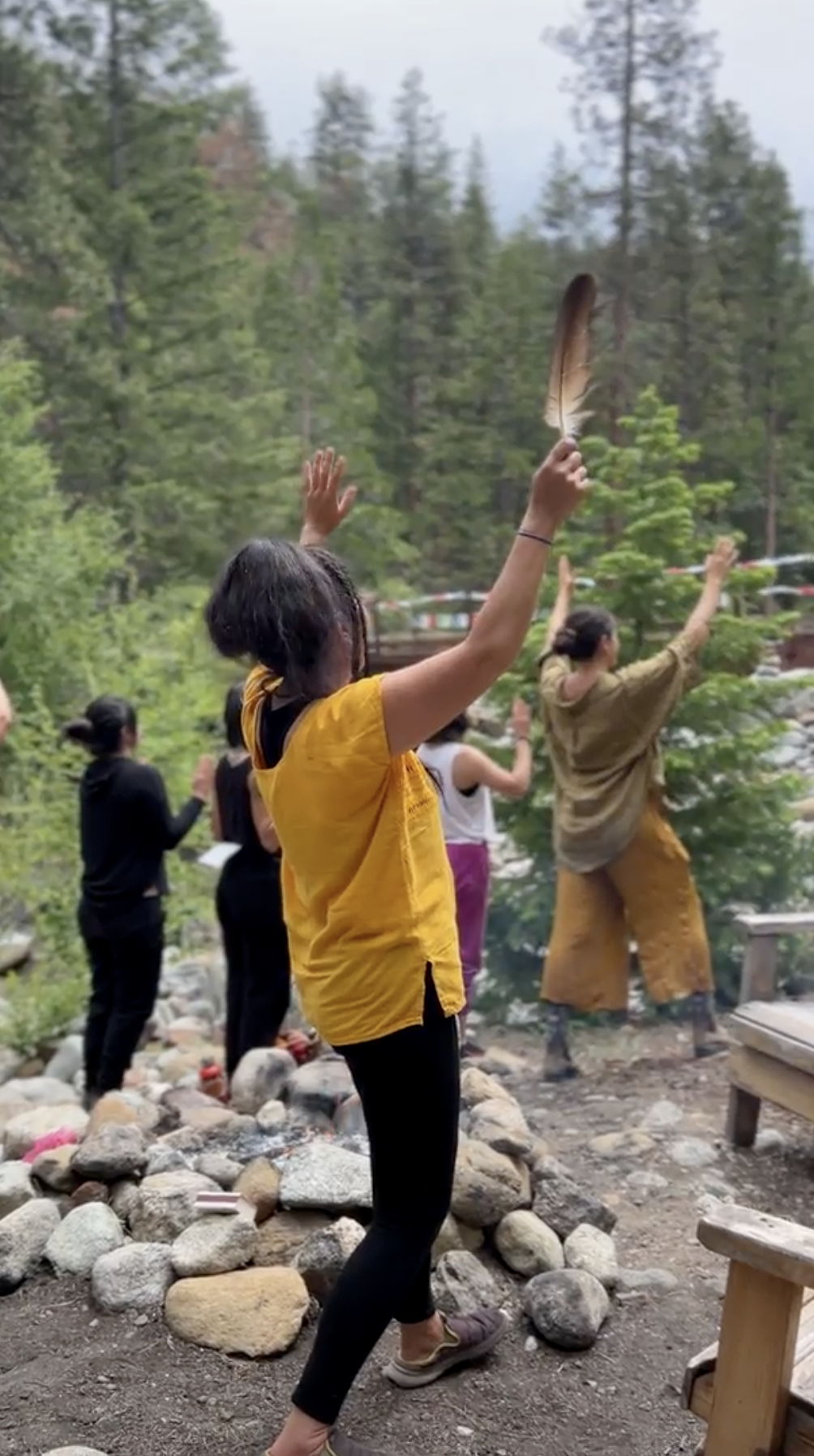 A group of people participating in a spiritual or meditation activity outdoors in a forested area, with one woman in a yellow shirt holding a large feather.