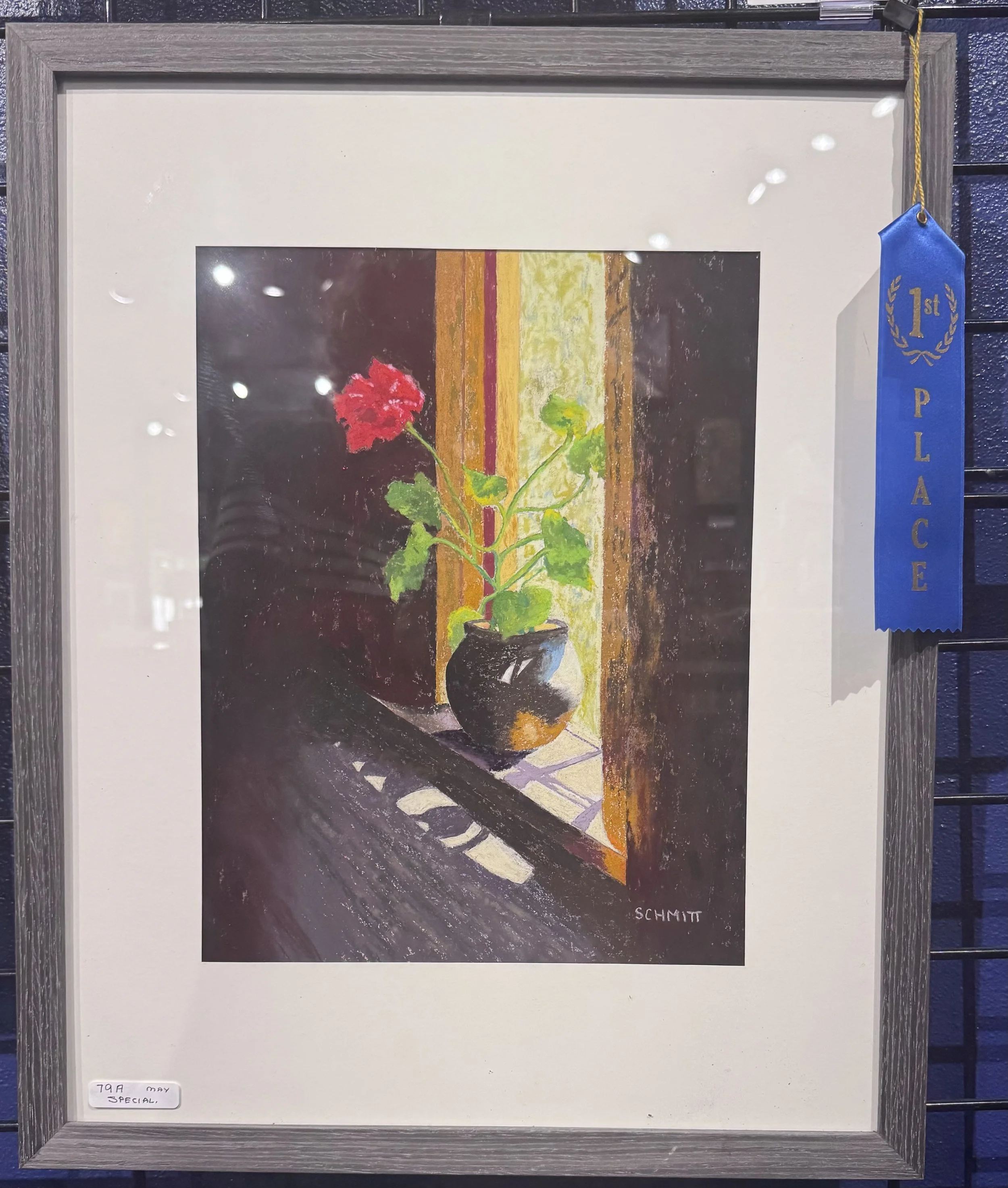 Framed artwork with a painting of a red flower in a pot, featuring vertical stripes in earthy tones in the background, signed by SCHMIT. A blue ribbon with gold lettering and a gold laurel wreath, indicating first place, is attached to the frame.
