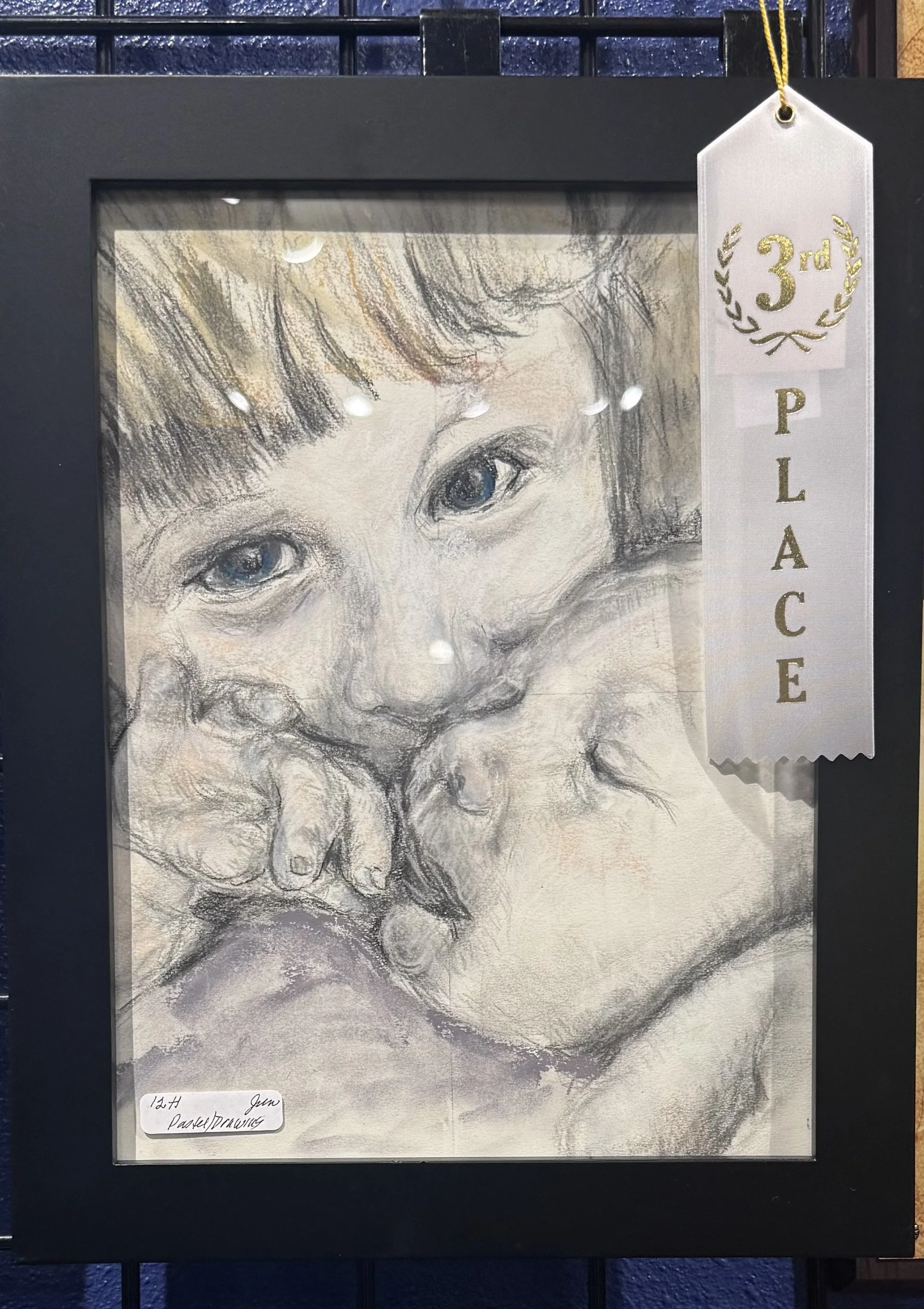 A framed pencil drawing of a young child's face, with large expressive eyes and curly hair, displayed with a white ribbon that reads '3rd PLACE'.