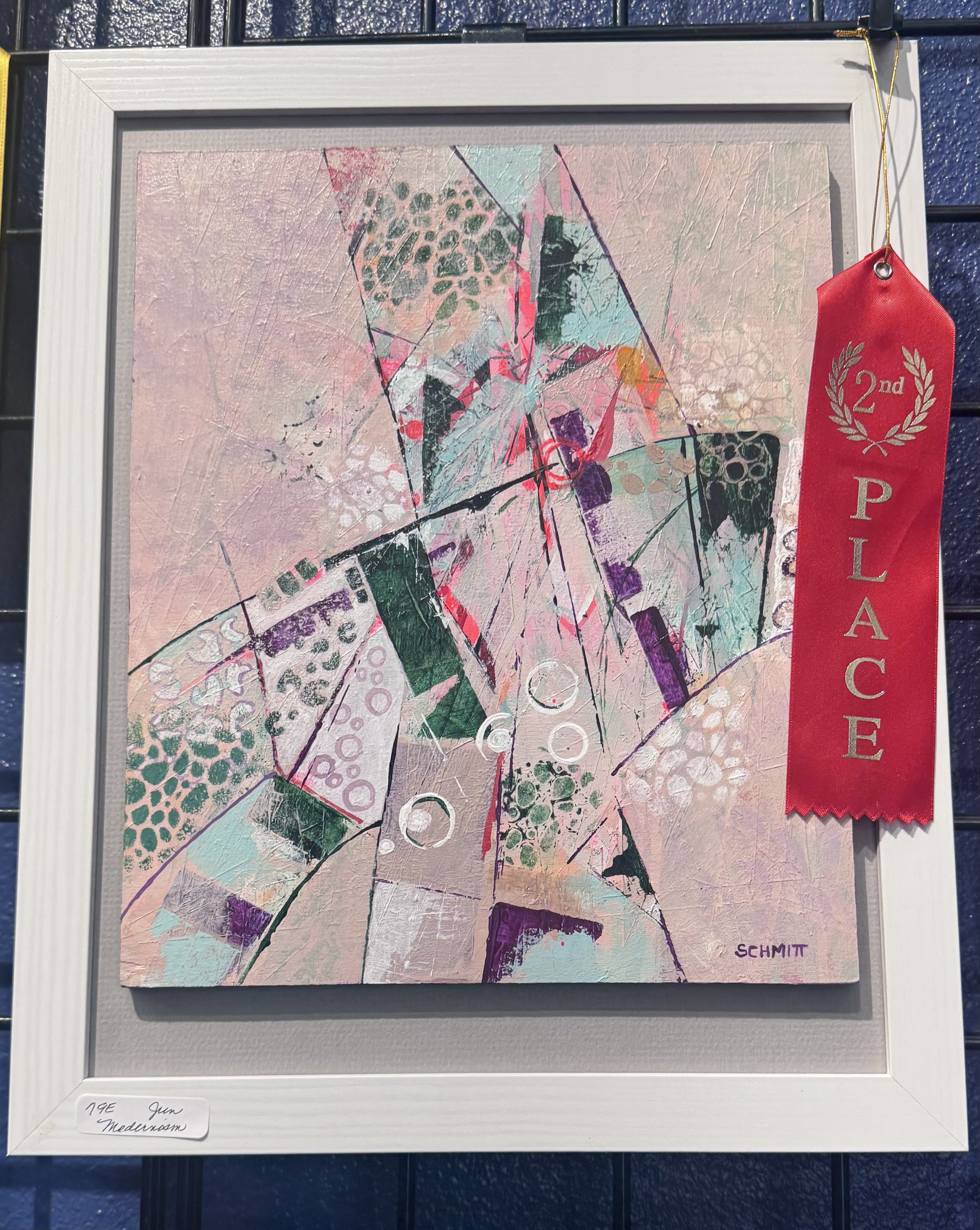 Abstract painting with geometric shapes and patterns in pastel pink, purple, teal, and black colors, framed with a white border and a red ribbon indicating second place. The artist's signature, 'SCHMIT,' is in the bottom right corner. A handwritten l