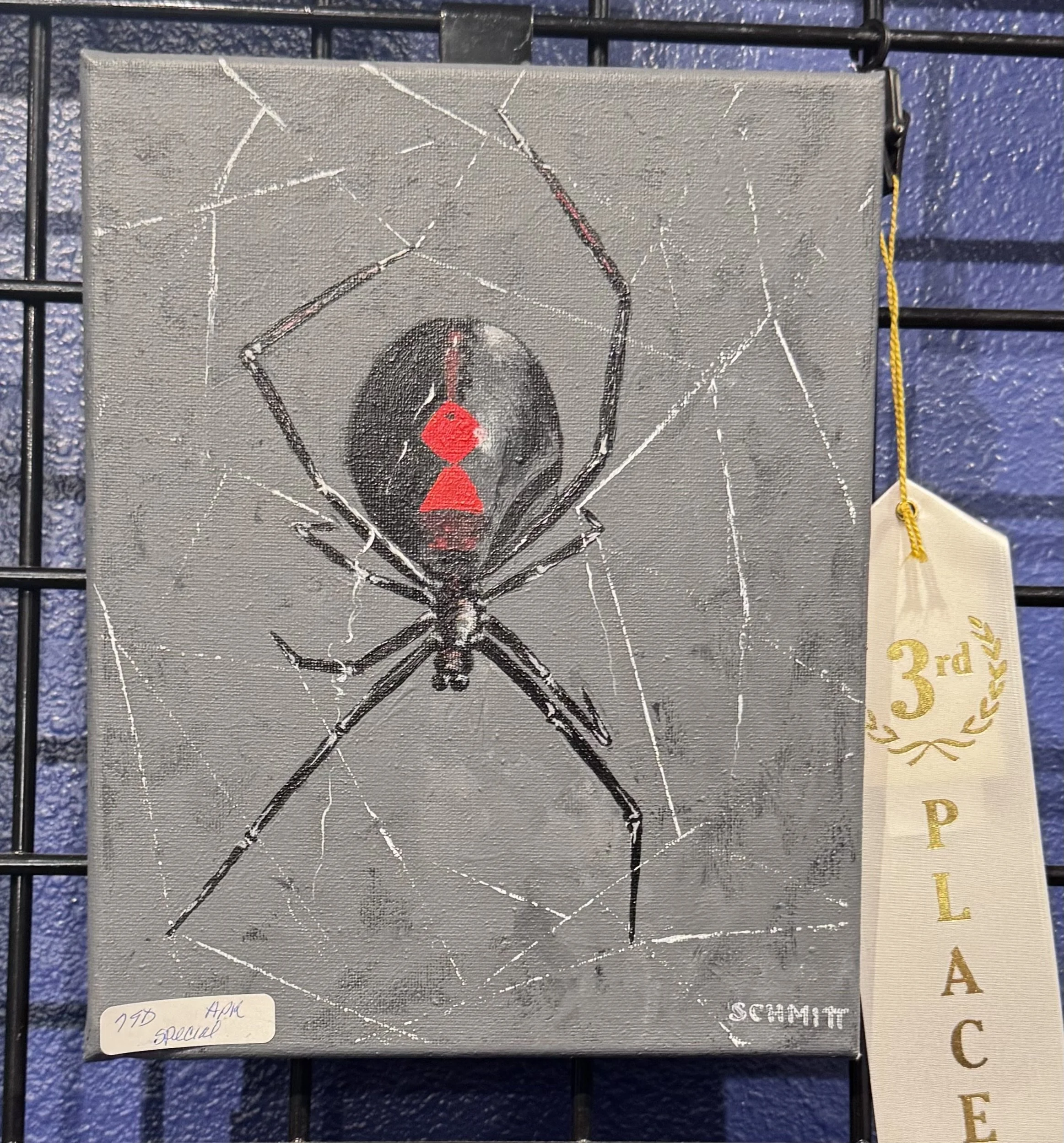 Painting of a spider with a human face surrounded by a web, displayed on a black grid with a gold third place ribbon beside it.