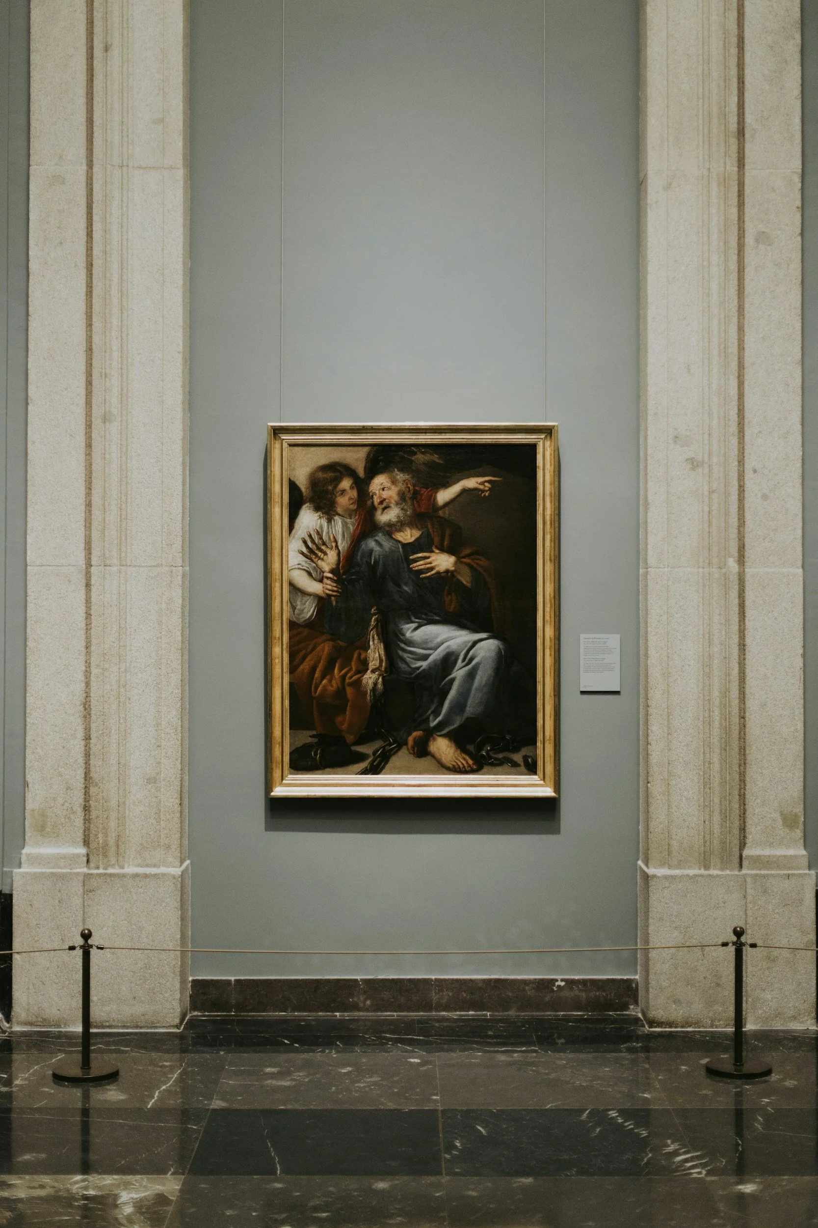 A painting of an older man with a beard, seated with chains around his feet, and a young girl whispering to him, with another man in the background, displayed in a museum gallery.