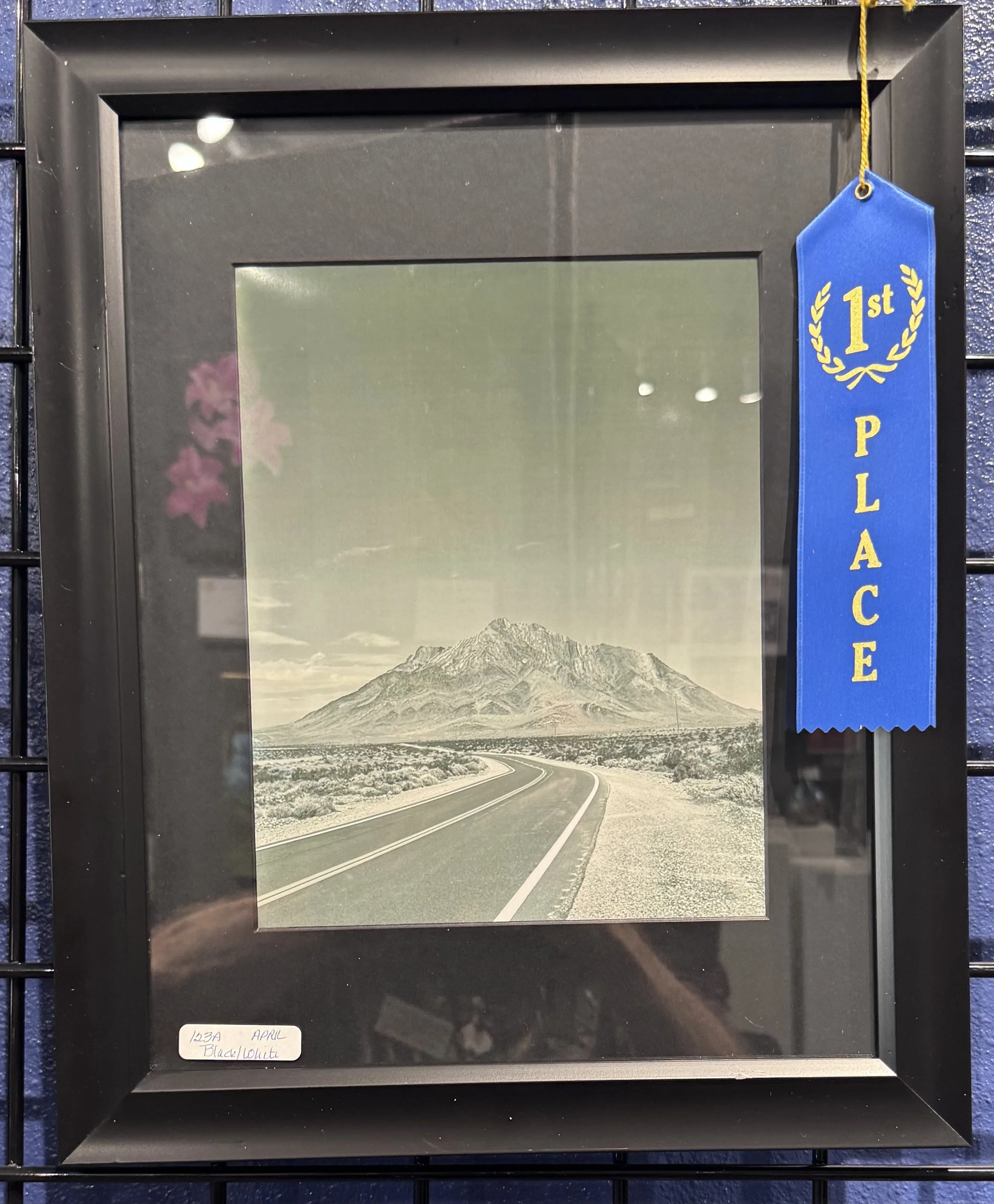 A framed black and white photograph of a mountain and a winding road, with a blue ribbon hanging on the top right corner that says '1st PLACE'. The frame is hung on a dark wall.