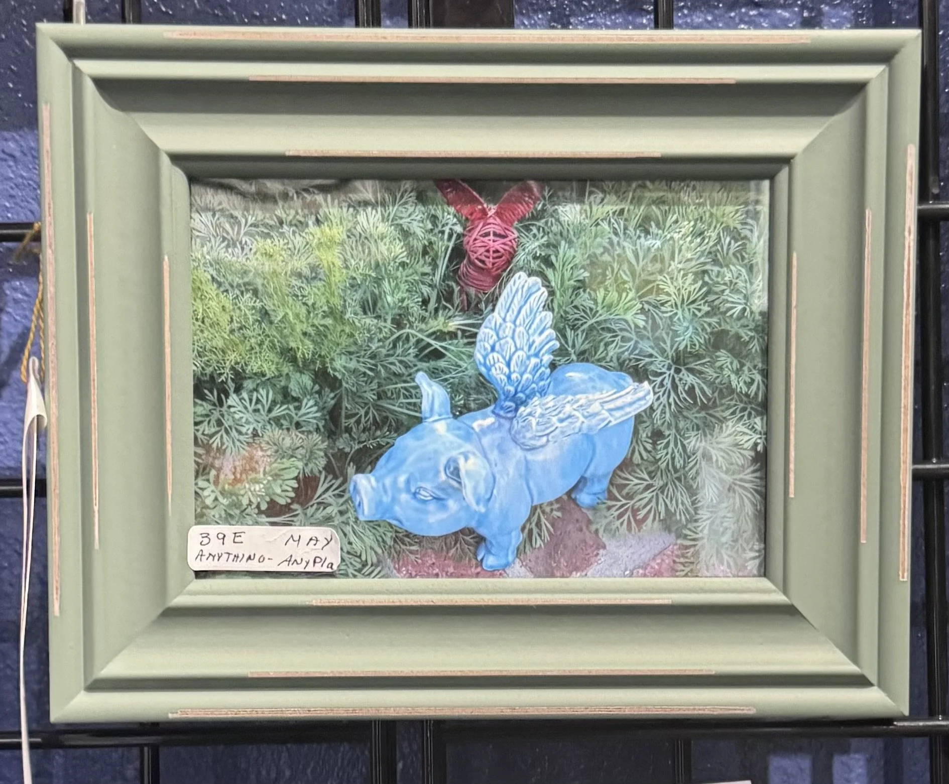 A framed artwork depicting a blue dove with a red ribbon tied around its neck, set against a background of lush green foliage and plants. There is a small handwritten note at the bottom left corner that reads: '39E May Anything-Anypla'.