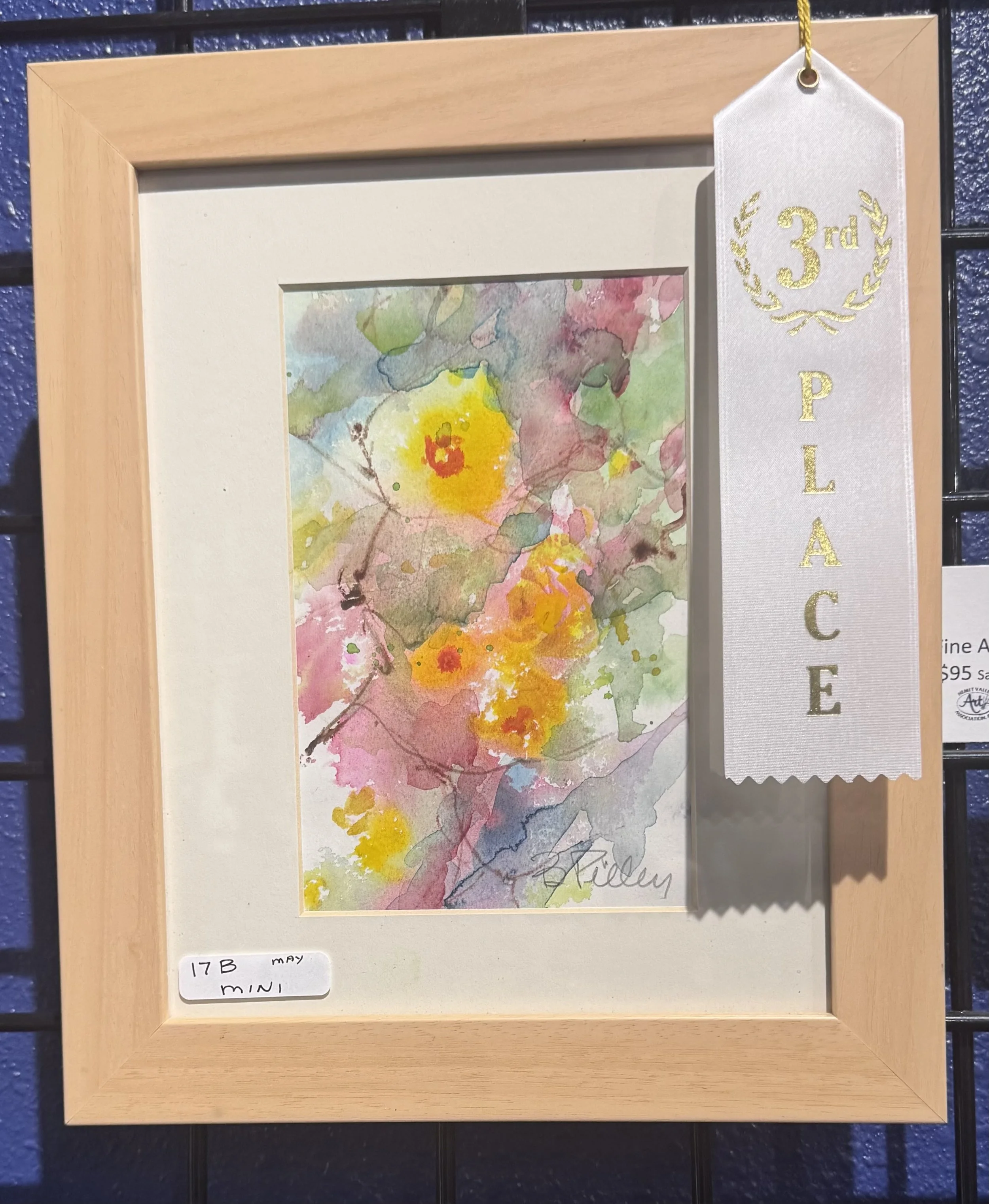 Watercolor painting of yellow and pink flowers with green and purple leaves, framed in a wooden frame with a white mat, displayed at an art exhibition.