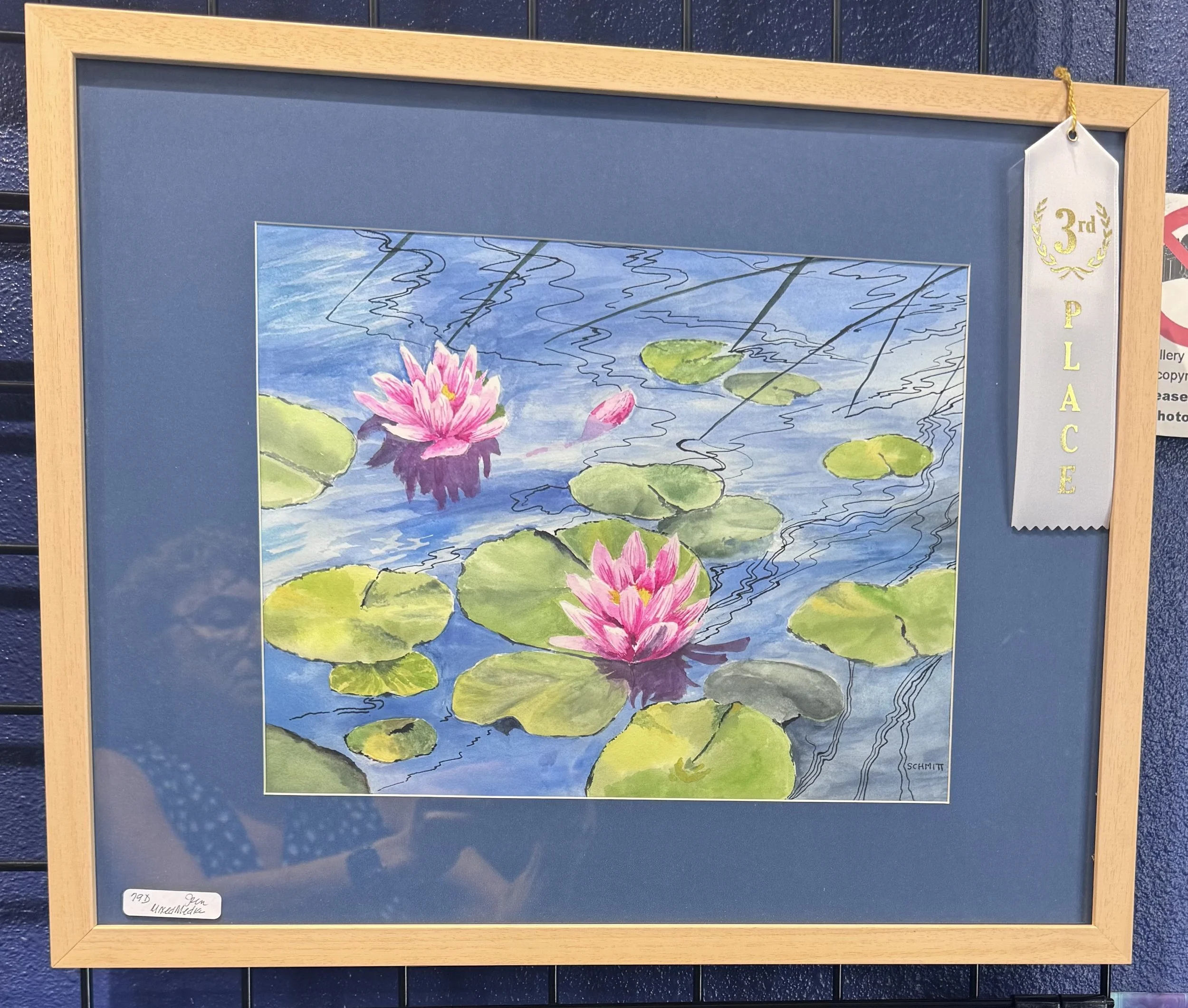 Water lily pond watercolor painting with pink water lilies and green lily pads. Gold and white third place ribbon hanging on the top right corner. The painting is framed in light wood. The artist's signature 'Schmitt' is visible in the bottom right c