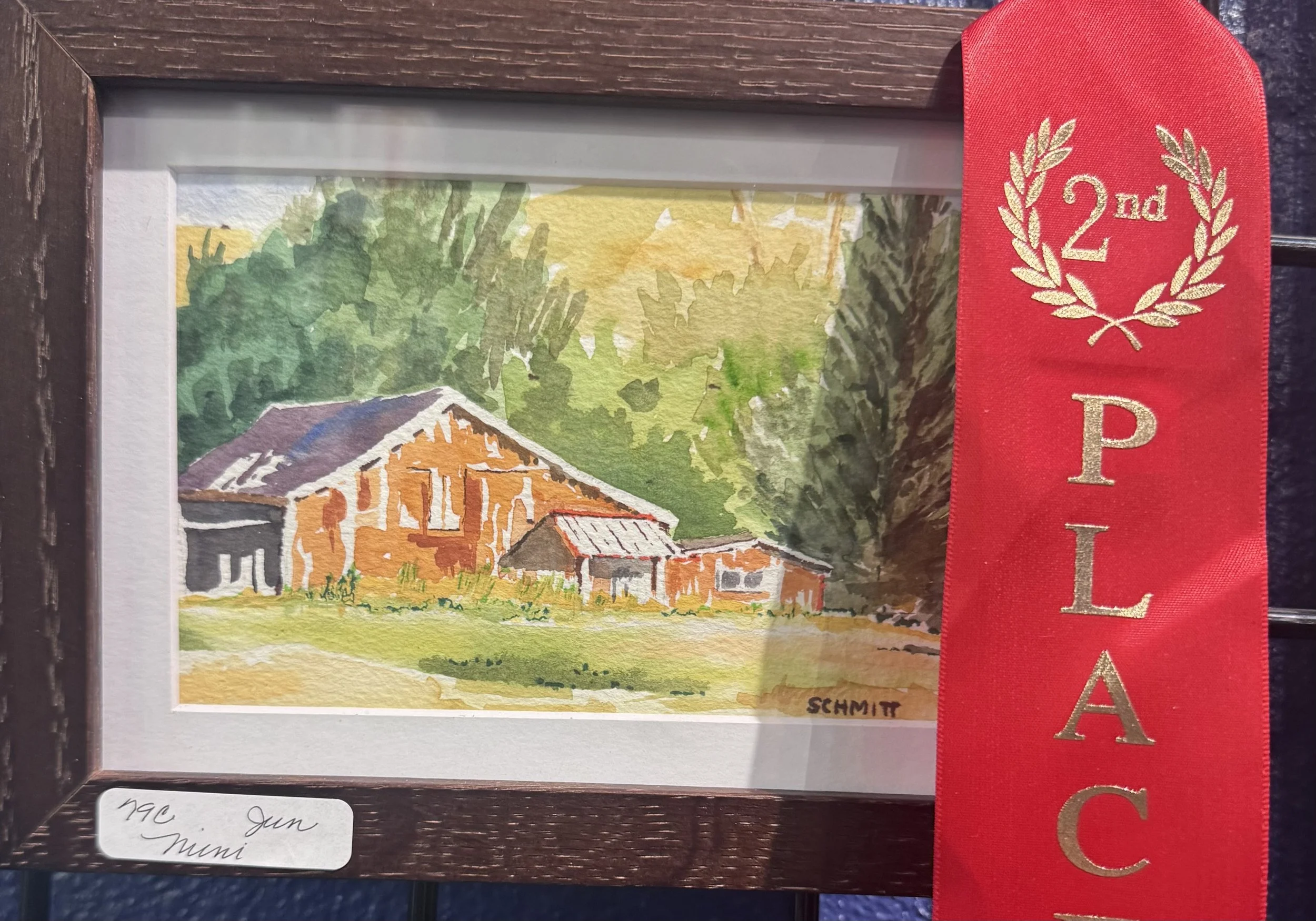 Watercolor painting of farm buildings with a barn and smaller structures, surrounded by green trees and hills, signed 'Schmitt'.