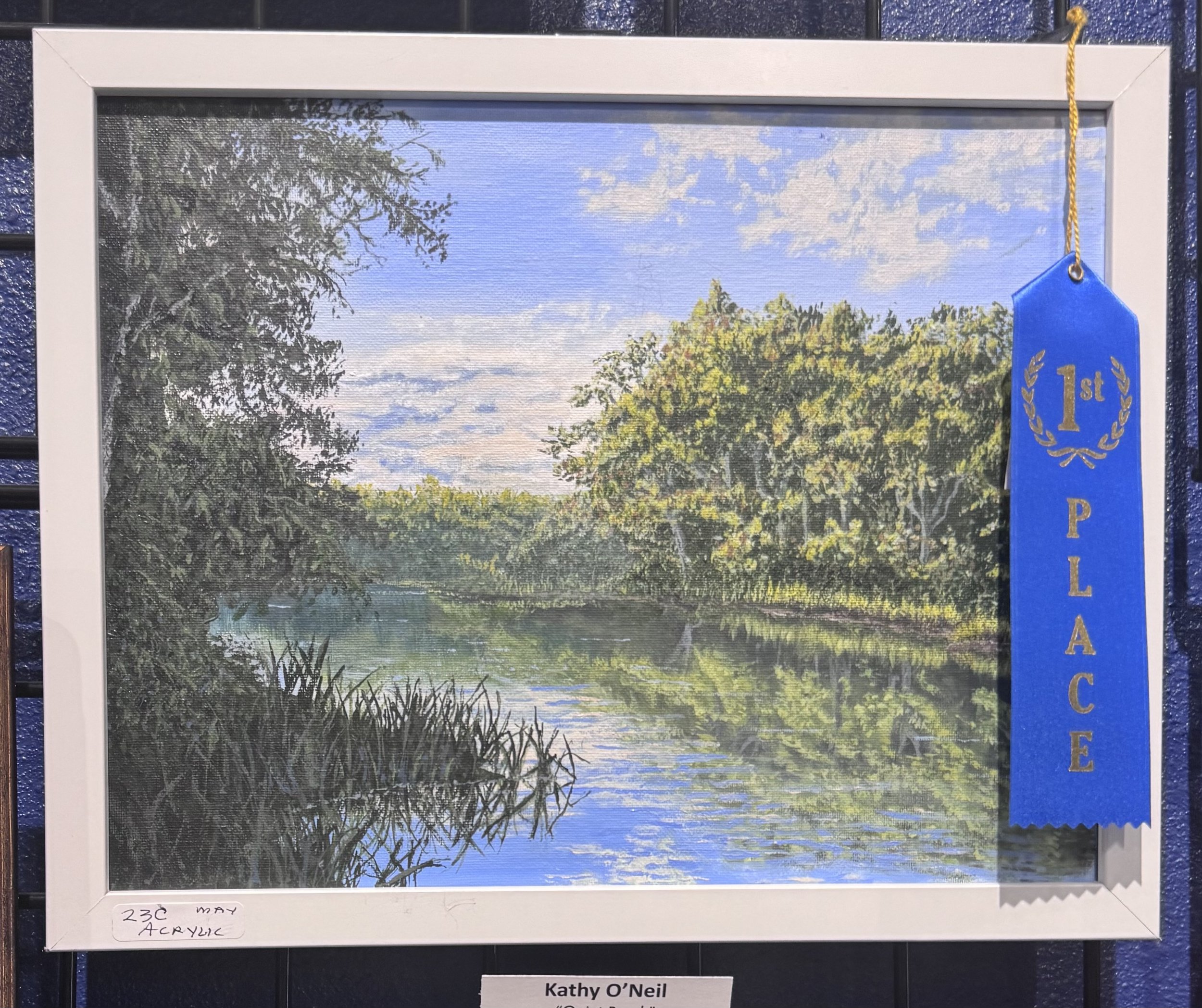 A landscape painting of a river with trees in the foreground and blue sky with clouds in the background. A blue ribbon labeled '1st' is attached to the top right corner of the painting.