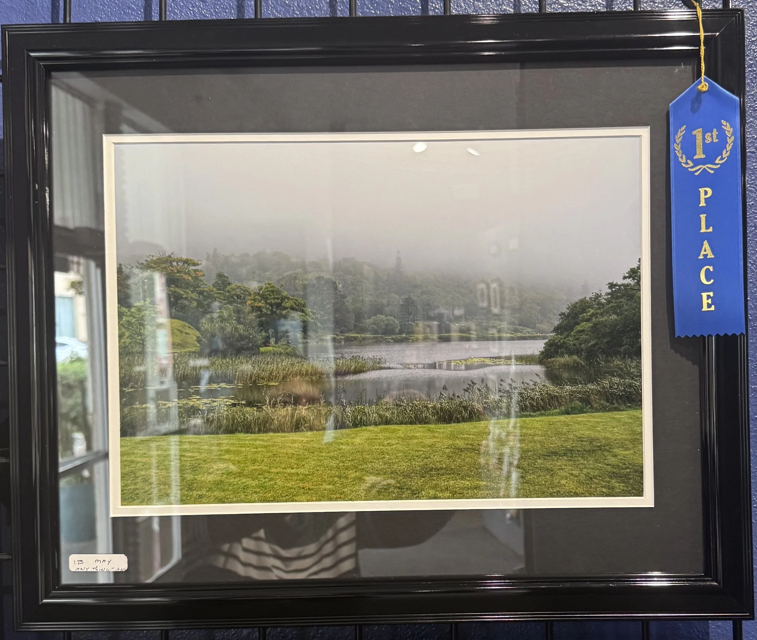 A framed mirror with a landscape scene of a river, trees, and fog. A blue ribbon with gold lettering reading "1st PLACE" hangs from the top right corner of the frame.