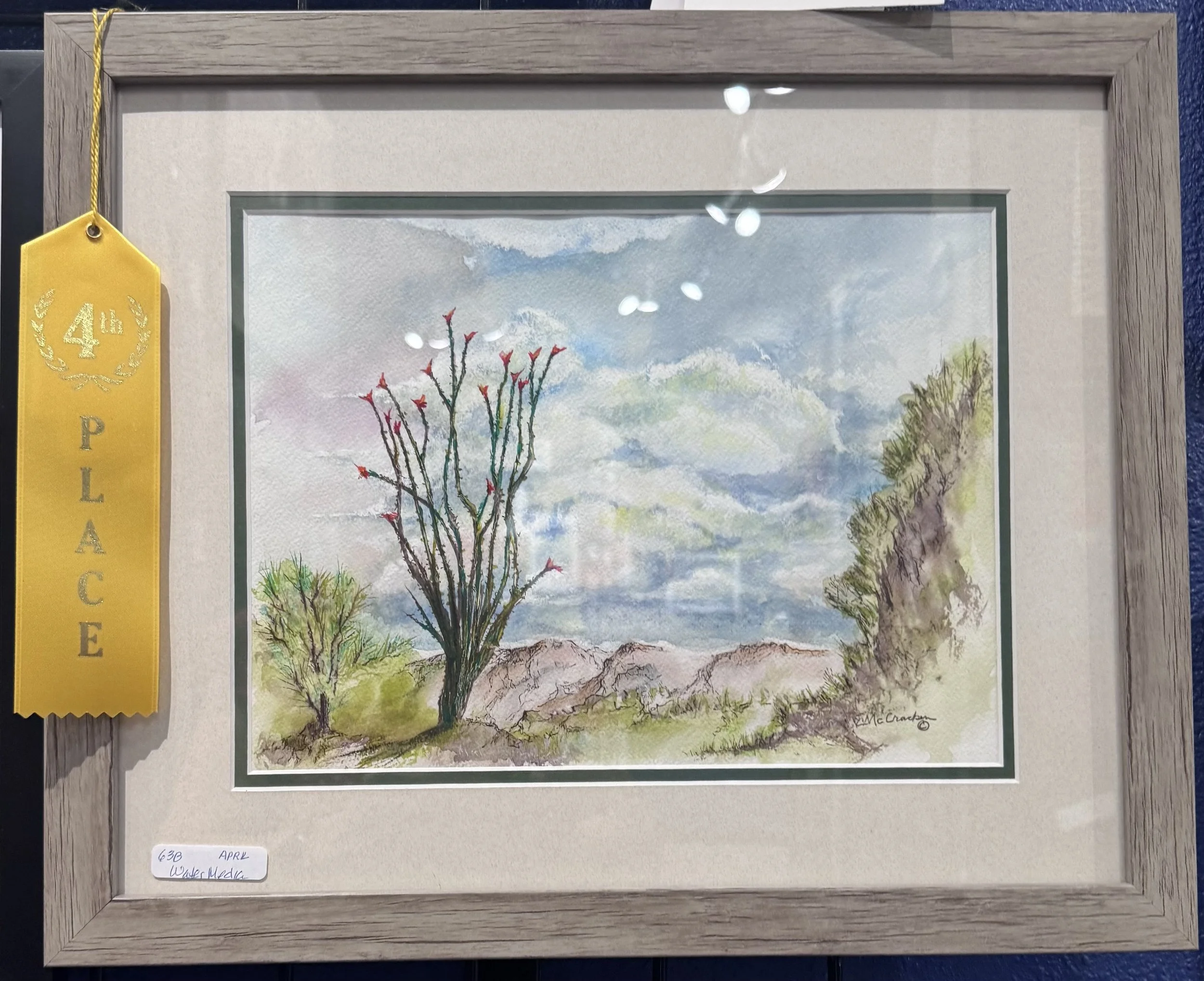 Watercolor landscape painting of a tree with red blossoms, mountains, and a cloudy sky, framed with a wooden frame, hanging on a wall with a yellow ribbon indicating 4th place.