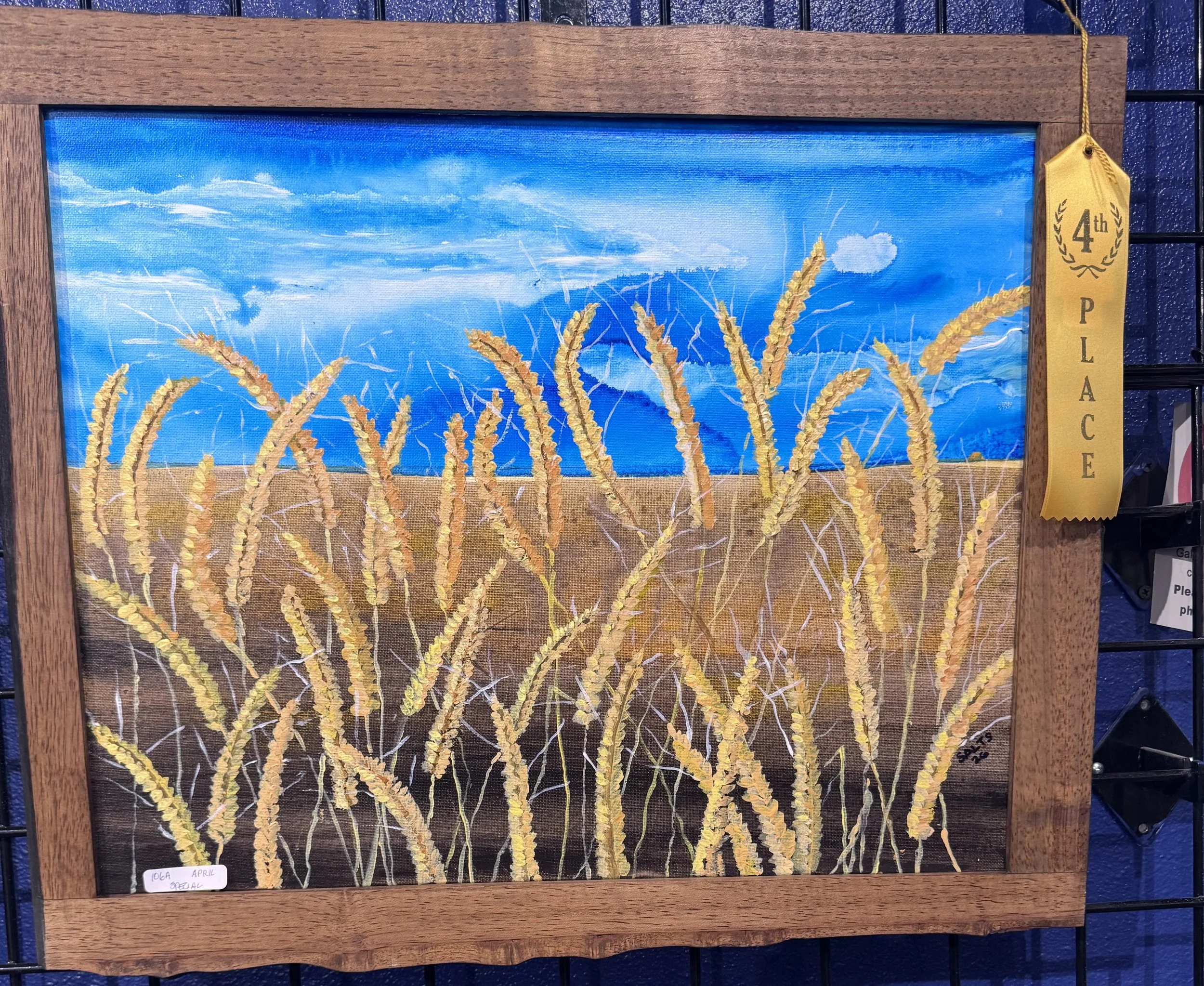 A painting of wheat field under a blue sky with clouds. The wheat stalks are golden yellow. There is a gold ribbon attached to the frame with text indicating 4th place.