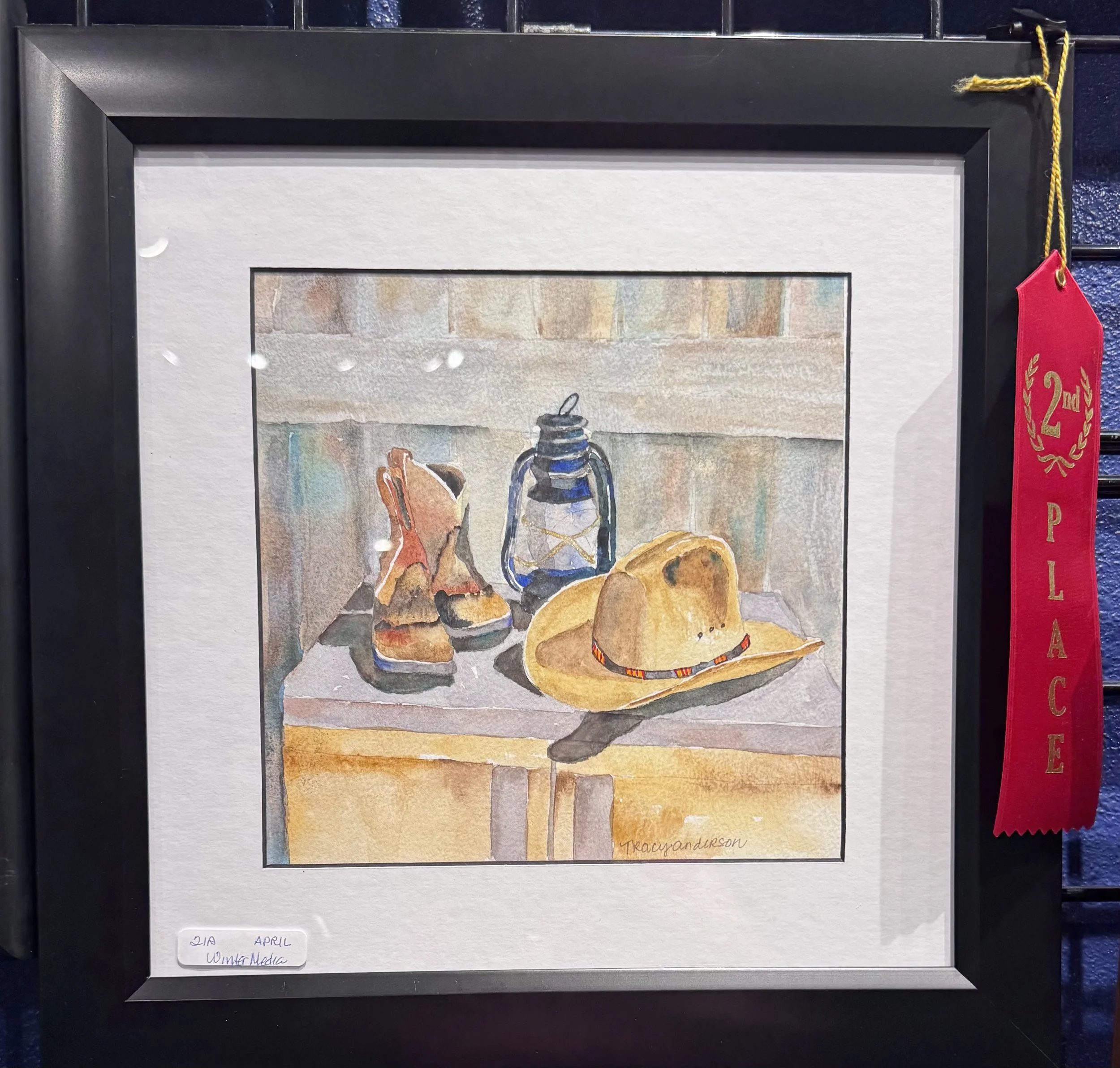 A framed watercolor painting of a still life arrangement with a brown pitcher, a blue thermos, and an orange object on a table, signed by Tracy Anderson.