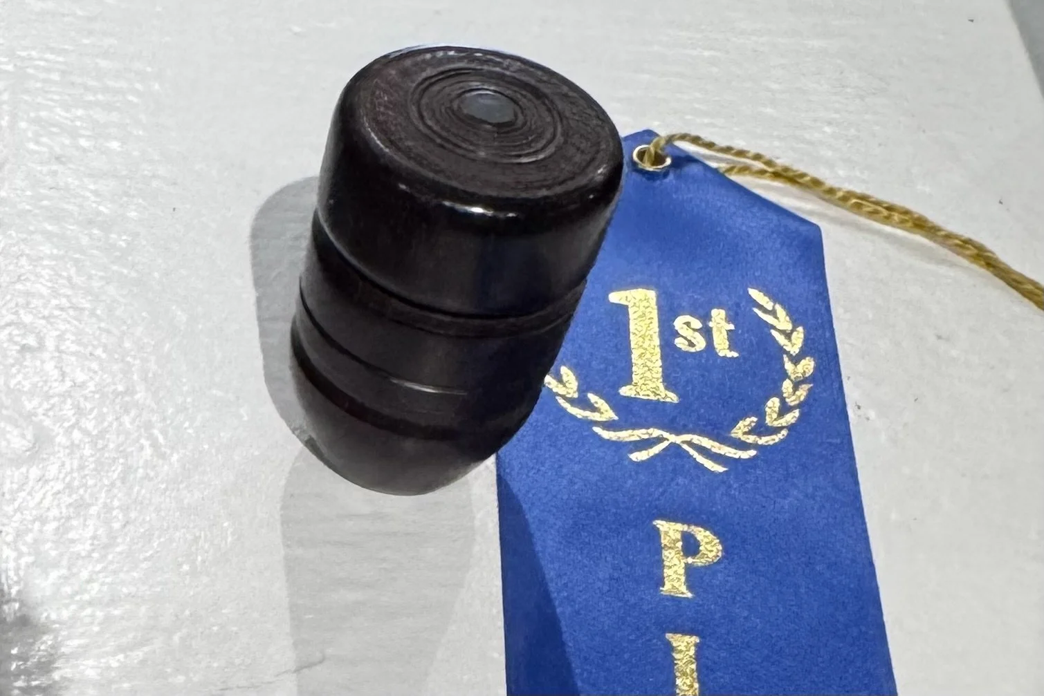 Close-up of a black barrel-shaped object. A blue ribbon with gold text and laurels, reading '1st P', is partially visible beside it. The object is on a light-colored surface.