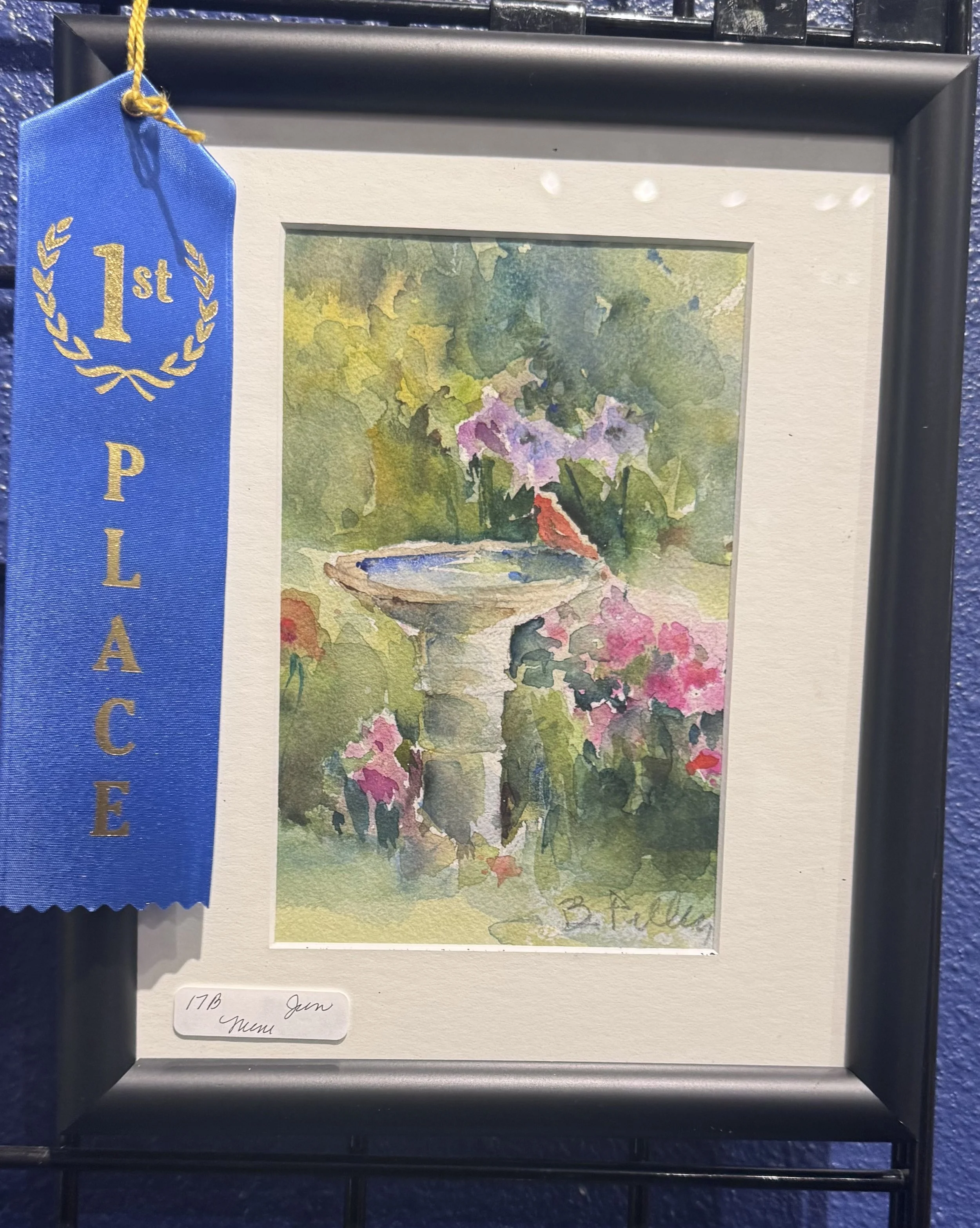 A framed watercolor painting of a garden scene with purple flowers, green foliage, and a small body of water, with a blue award ribbon attached to the left side of the frame.