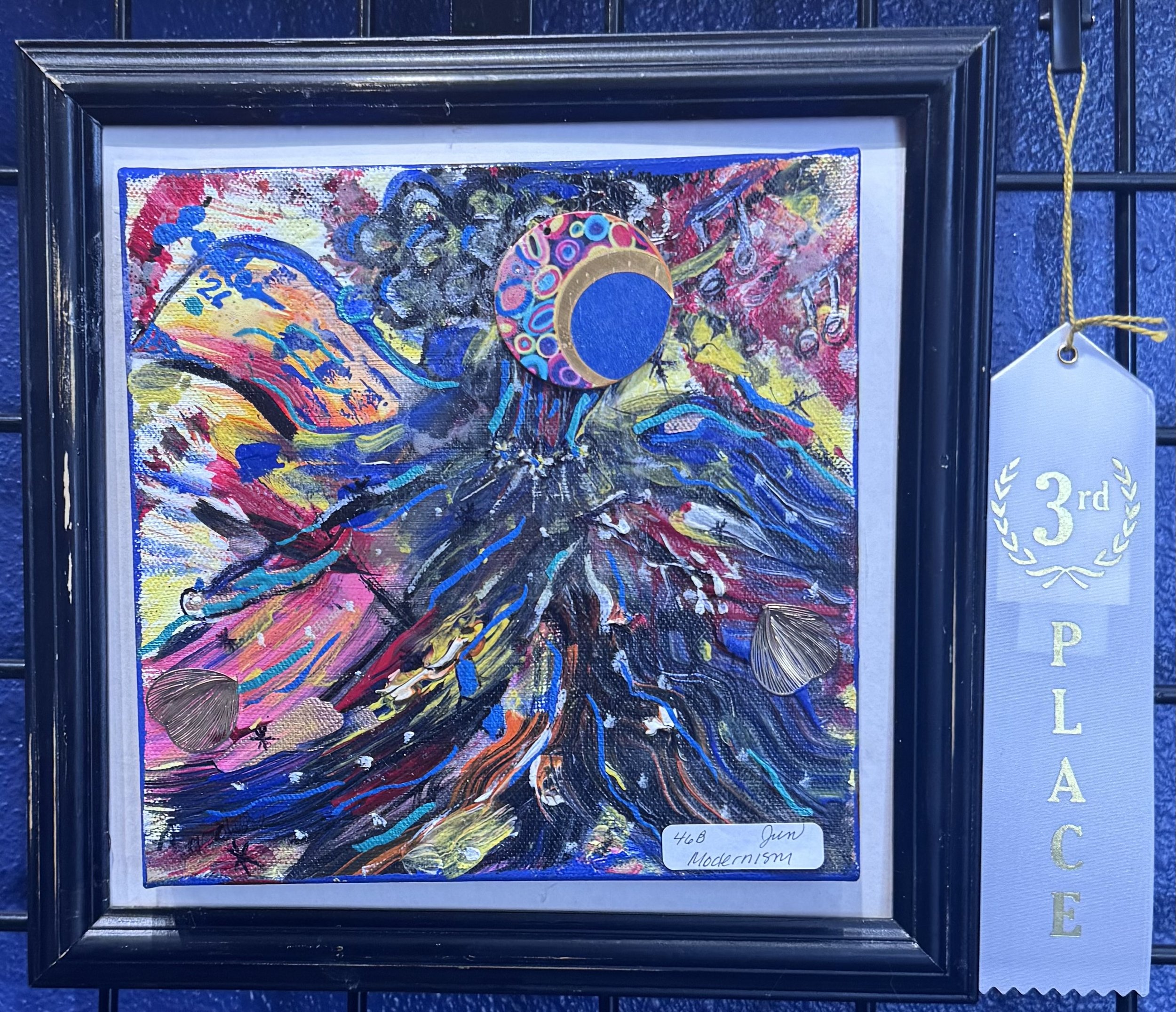 Abstract painting with vibrant colors, including blue, pink, yellow, and black, framed and displayed on a black rail. It has a circular decorative element with colorful circles and gold accents, and a small white label at the bottom right corner indi