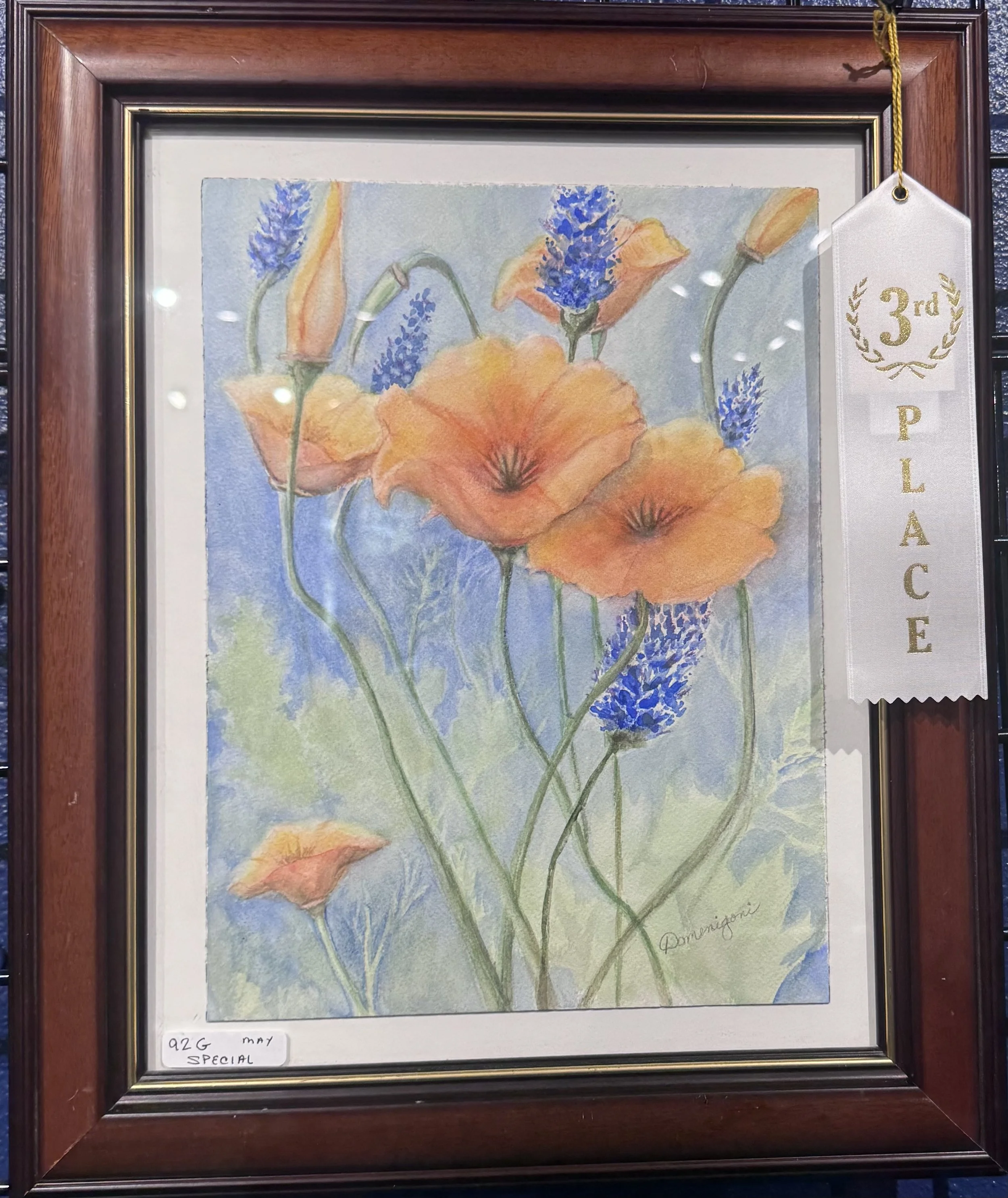Framed watercolor painting of orange poppies with blue flowers, signed by the artist. A white ribbon with gold lettering and laurel design indicating third place is attached to the frame's top right corner.