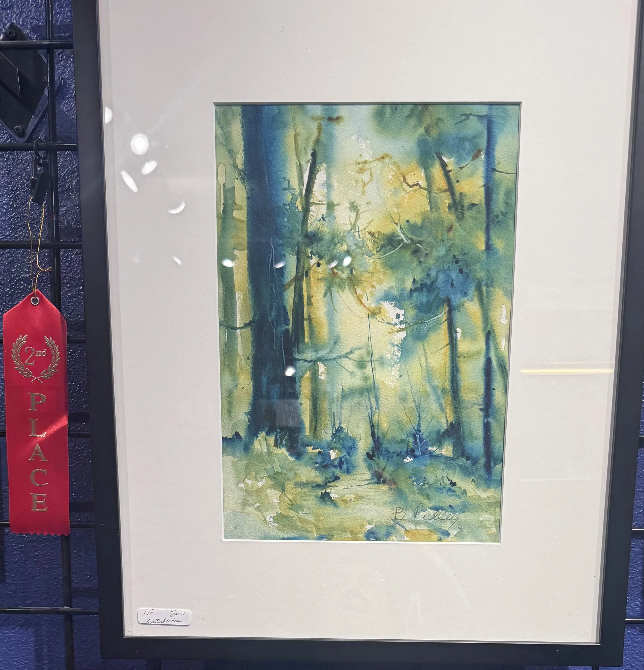 A watercolor landscape painting with trees and a body of water, displayed on a wall with a white mat and black frame. There is a red ribbon award on the left side indicating second place.