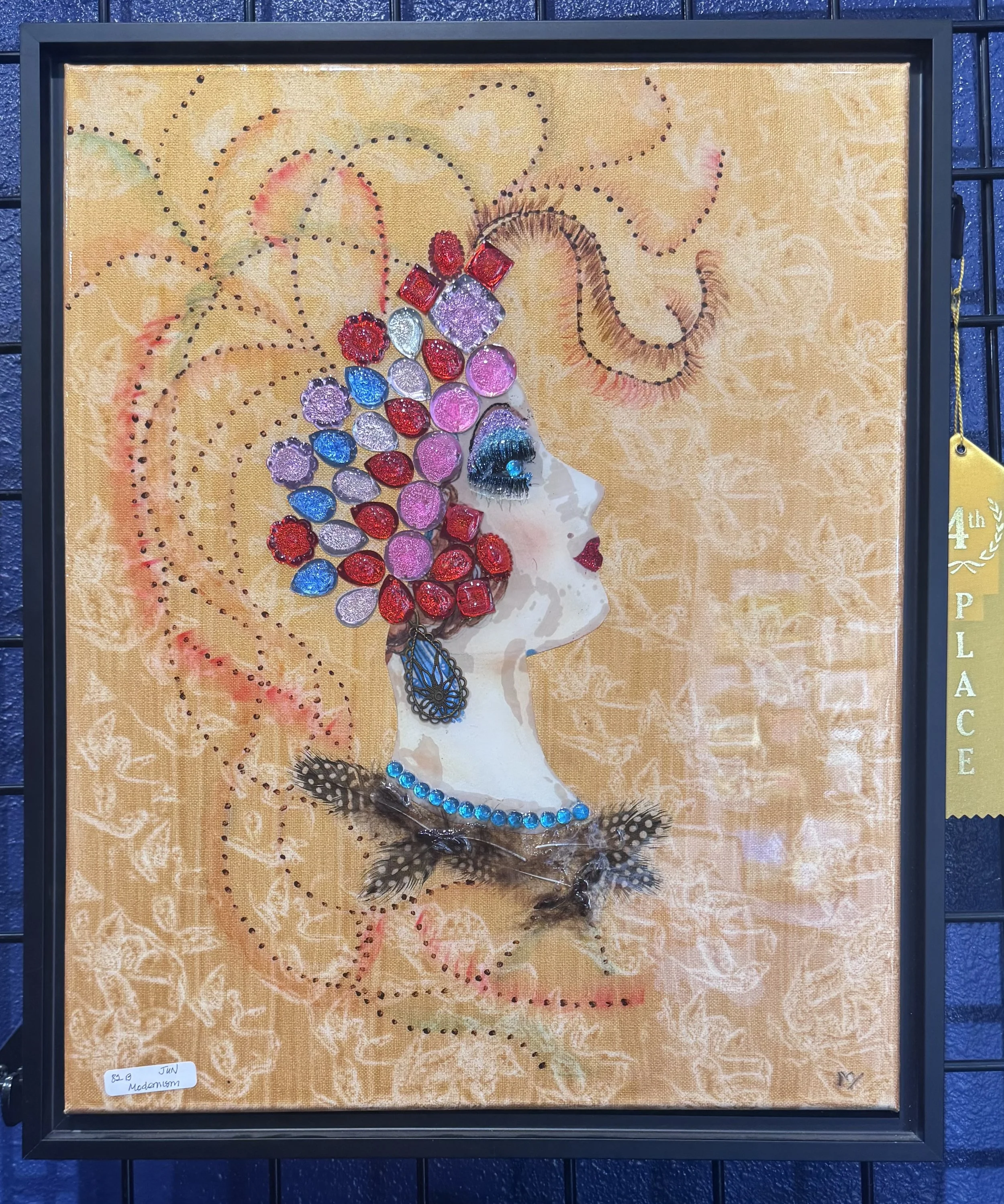 Artwork of a mermaid made with colorful gemstones and jewelry set against a golden background with decorative swirls and dots, framed in black.