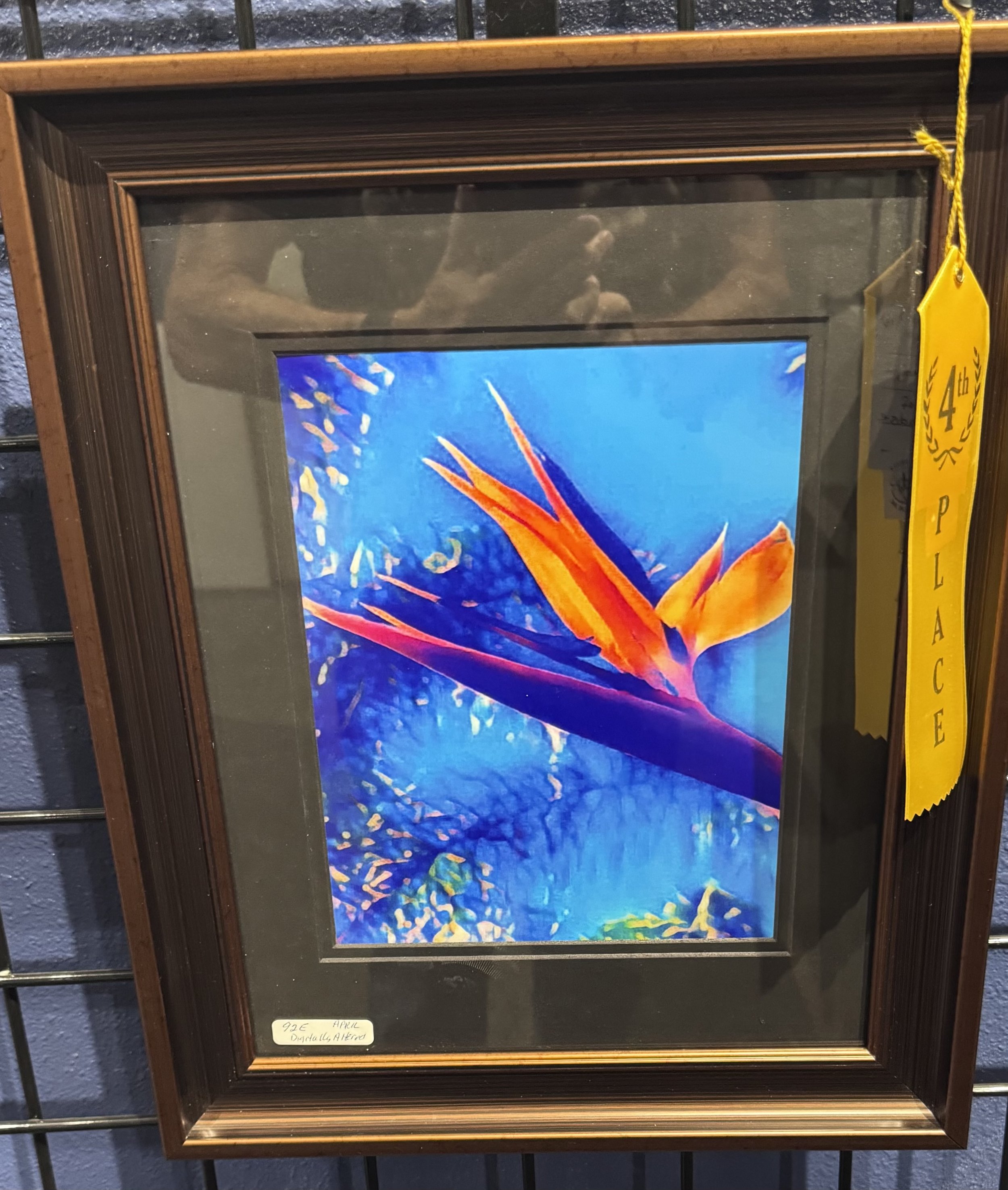 Framed colorful abstract painting of a bird of paradise flower with vibrant orange and yellow petals and a purple and green stem, set against a blue background, at an art auction or gallery, with a yellow ribbon indicating 4th place.