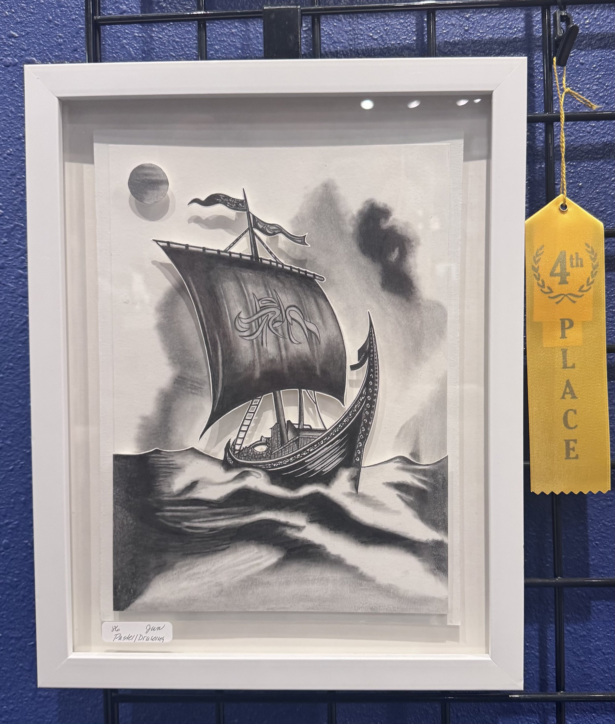 Black and white drawing of a Viking ship on choppy seas with a large sun or moon in the sky. The ship has a dragon figurehead and a flag at the stern. The drawing is framed and part of an exhibit labeled as '6 Gun Pastel/Drawing,' with a yellow ribbo