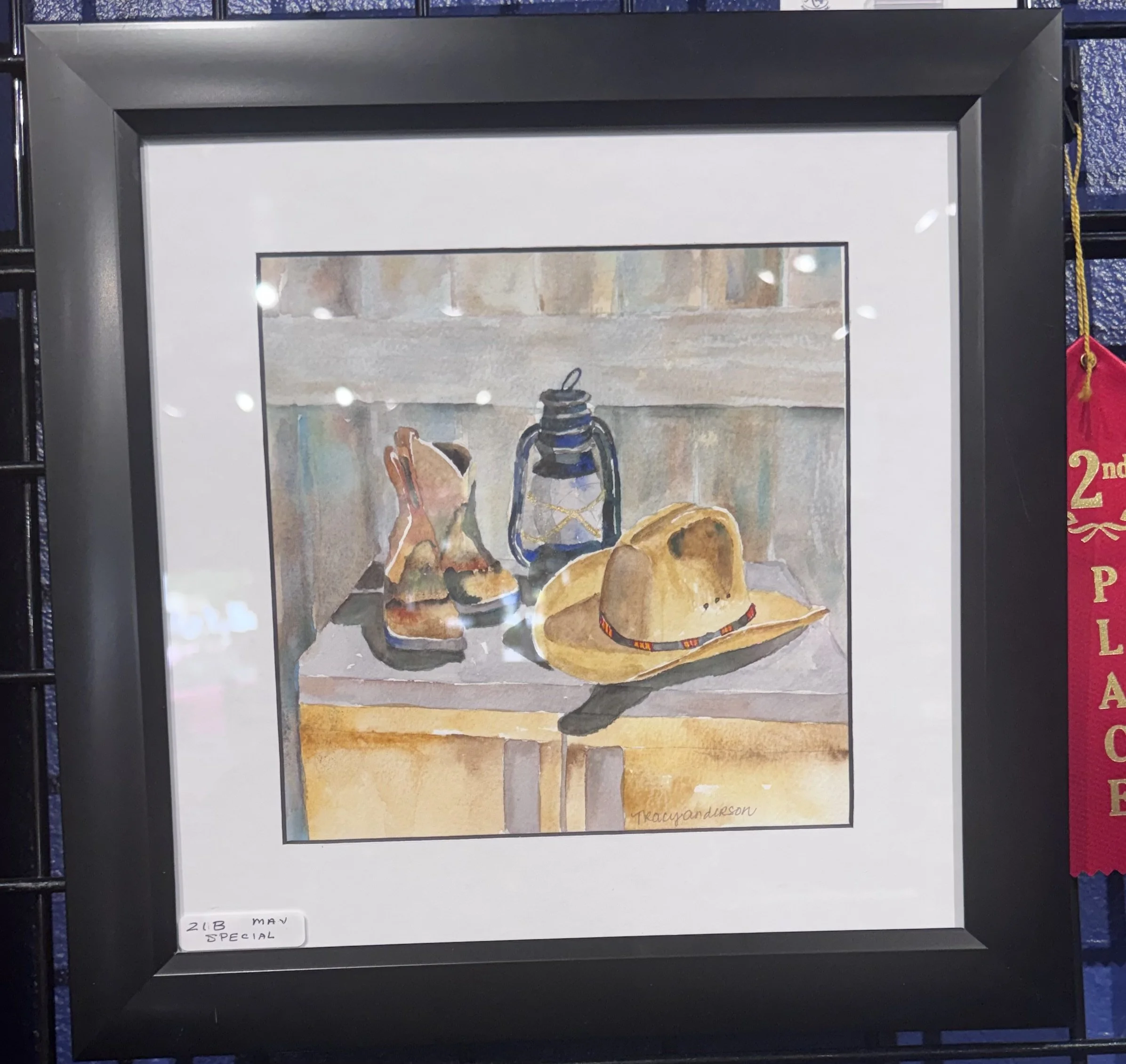 Framed watercolor painting of cowboy boots, a woven bag, and a cowboy hat arranged on a surface, with a small white paper label reading '2 LB May Special' in the bottom left corner.