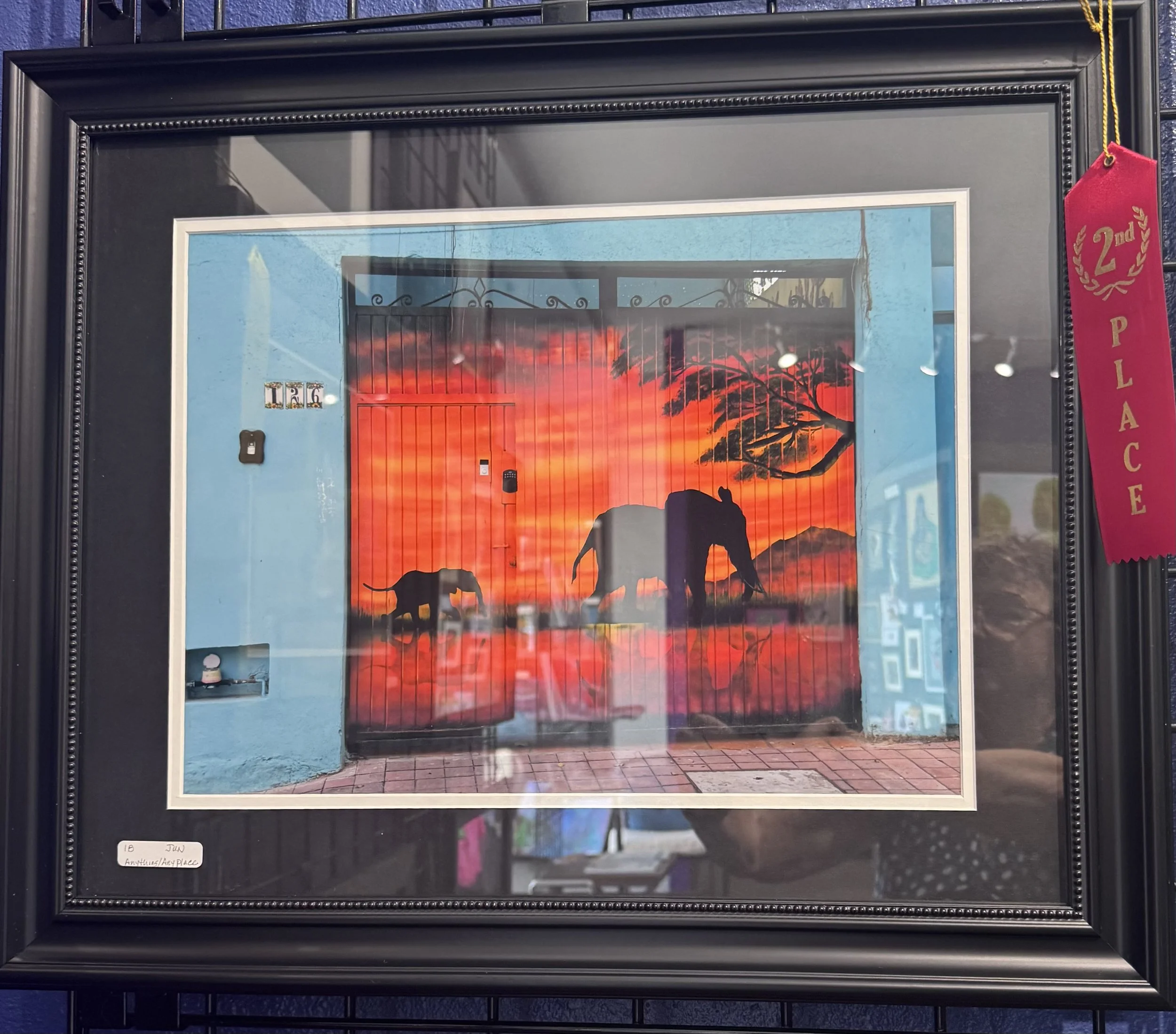 Framed artwork featuring a sunset or sunrise with silhouettes of a giraffe, tree, and mountain scene. Red, orange, and black colors dominate the image.