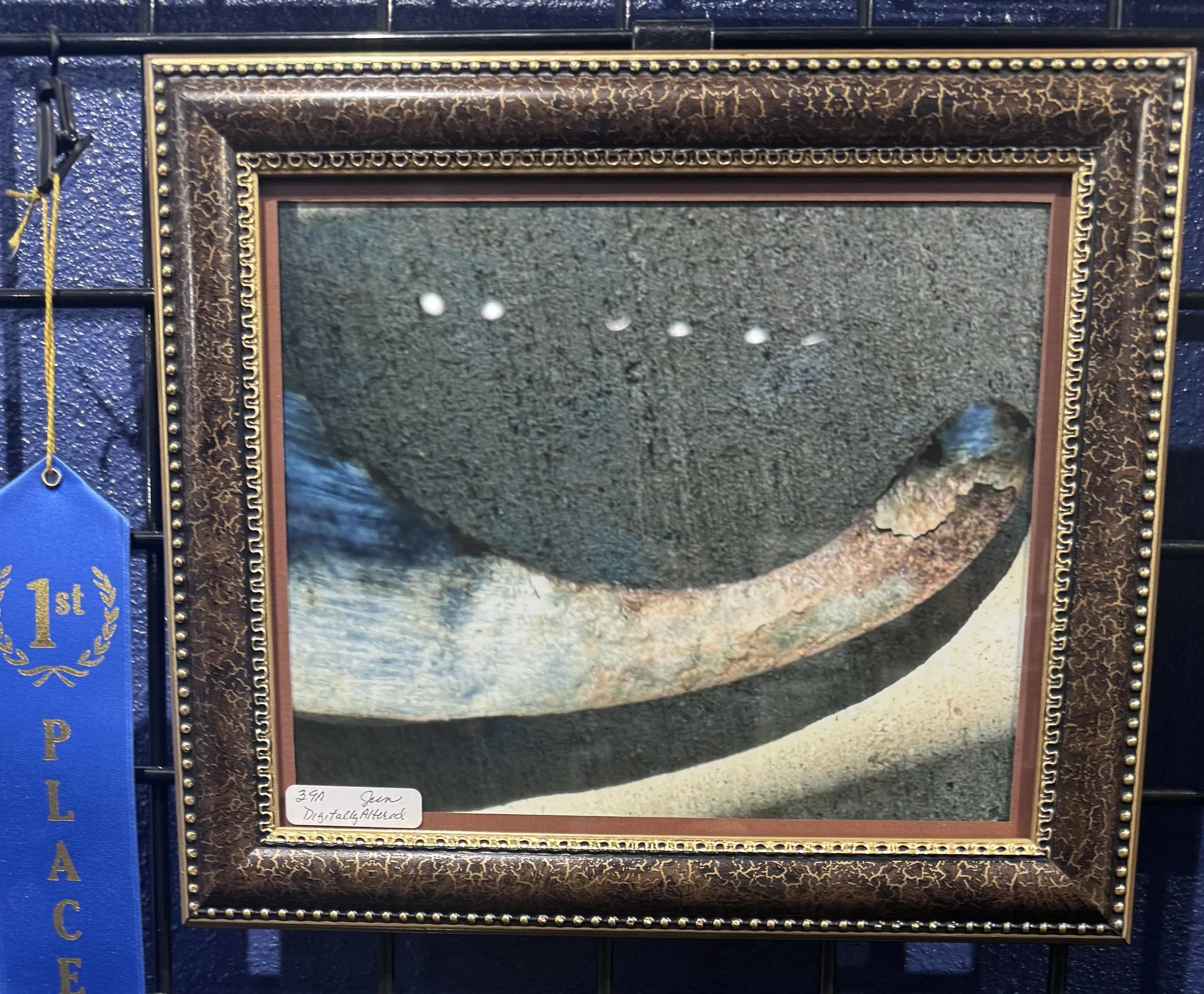 A framed gemstone resembling a large, rounded, brownish-green mineral with a rough surface, displayed on a textured blue background.