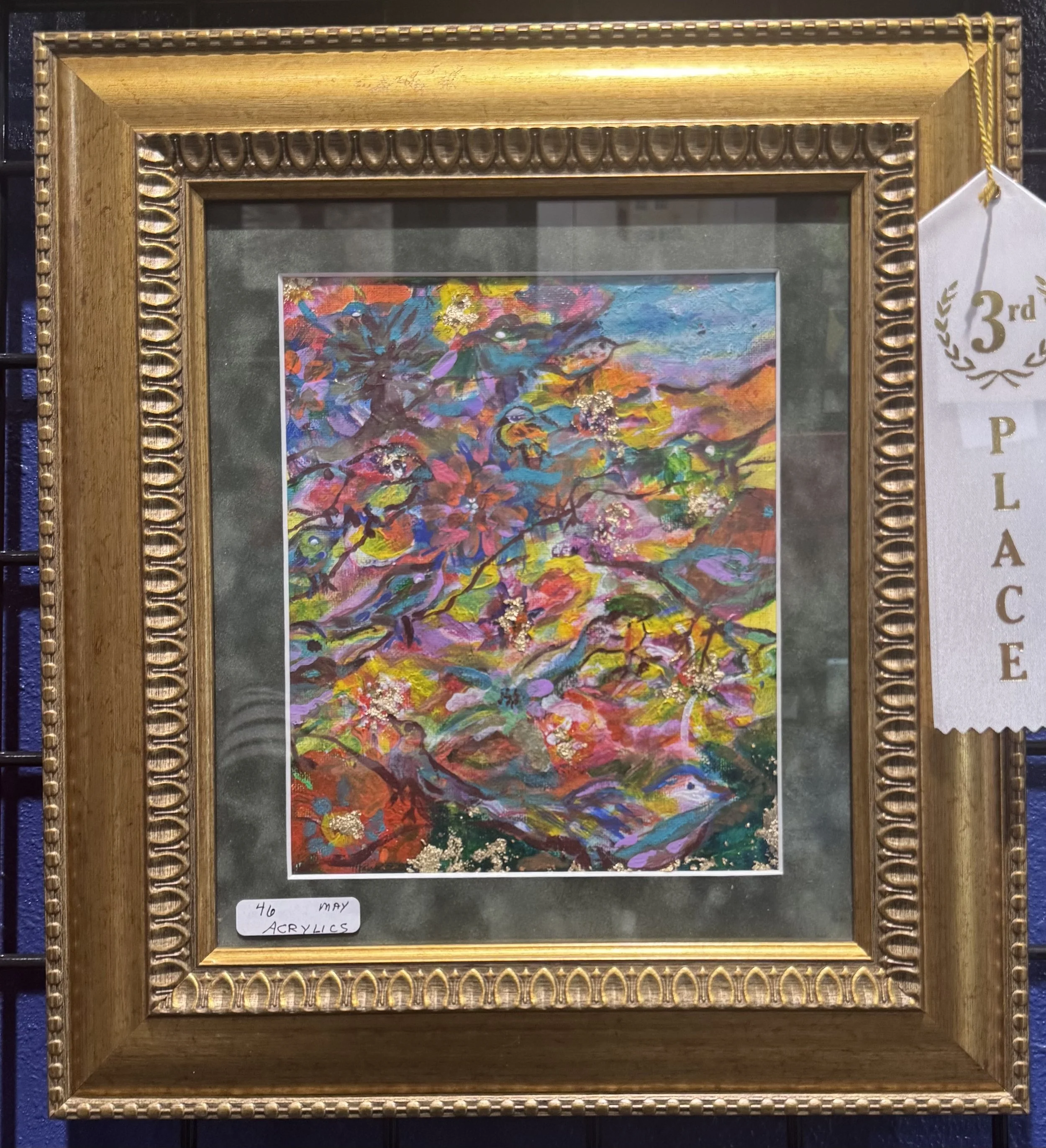 Close-up of a framed colorful abstract painting with pink, purple, blue, green, yellow, and orange colors on a black background. A white label at the bottom left corner reads '40 May Acrylics'. To the right, a white ribbon with gold text indicating t