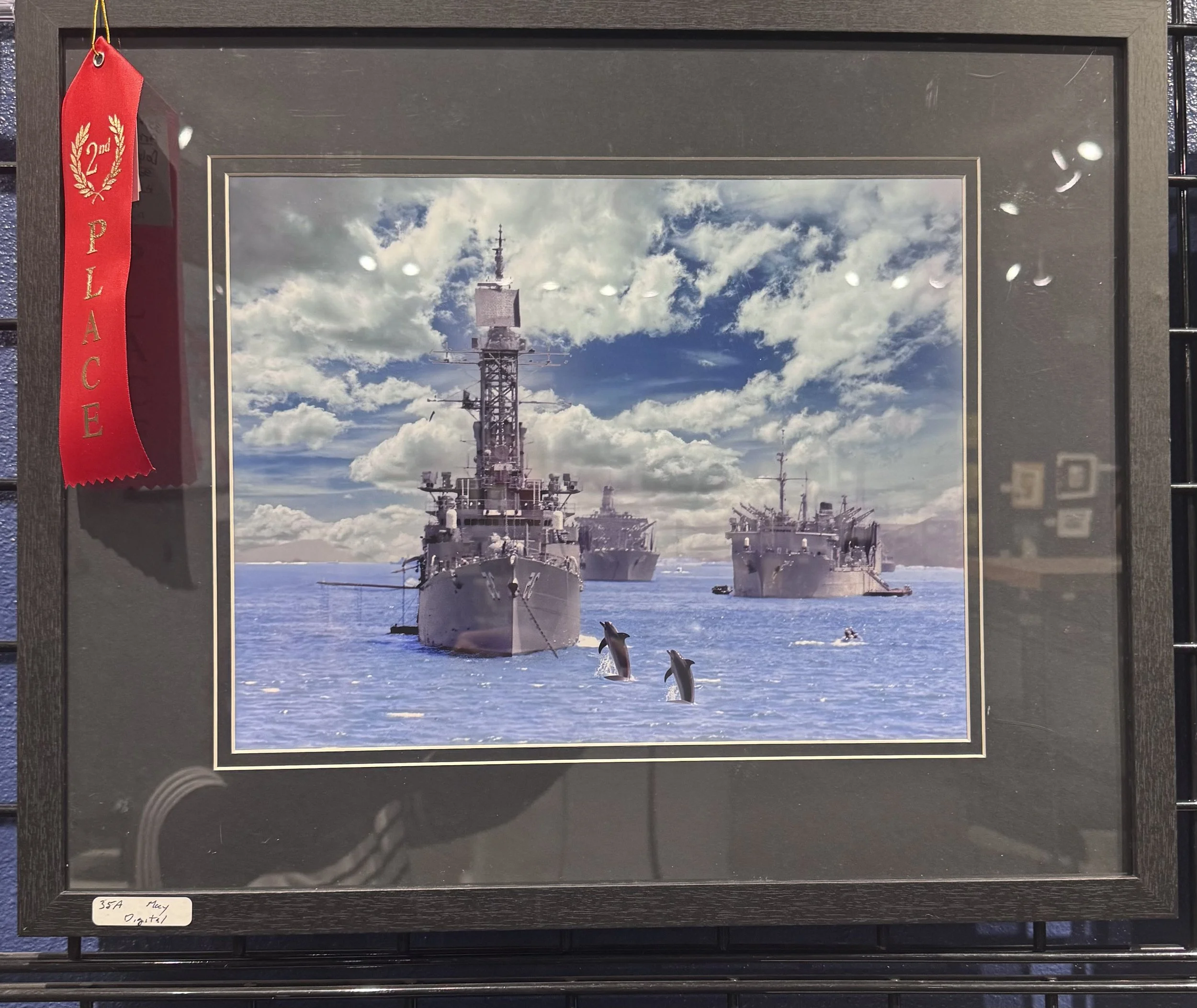 Photograph of three military ships in the ocean with a partly cloudy sky in the background, taken from a framed picture on display.