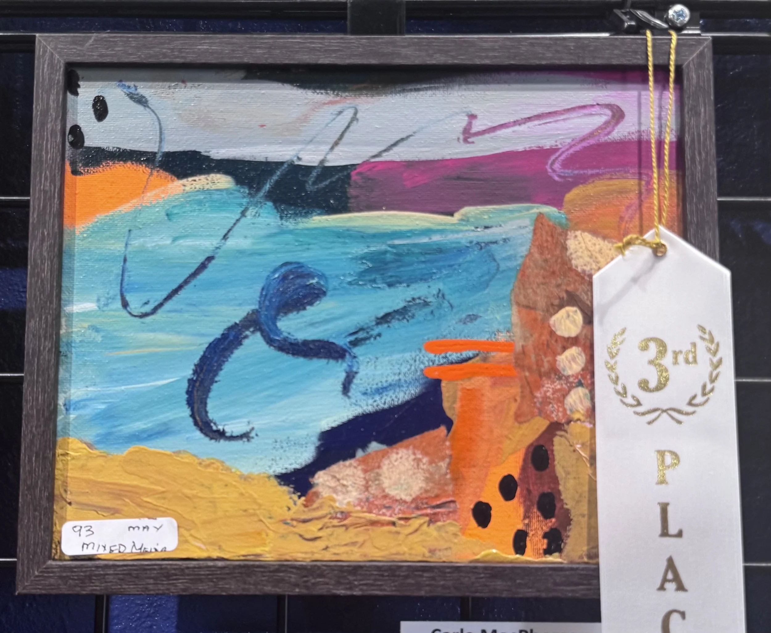 Close-up of an abstract colorful painting framed with a dark wood border, with a small white label attached to the bottom right corner of the frame.