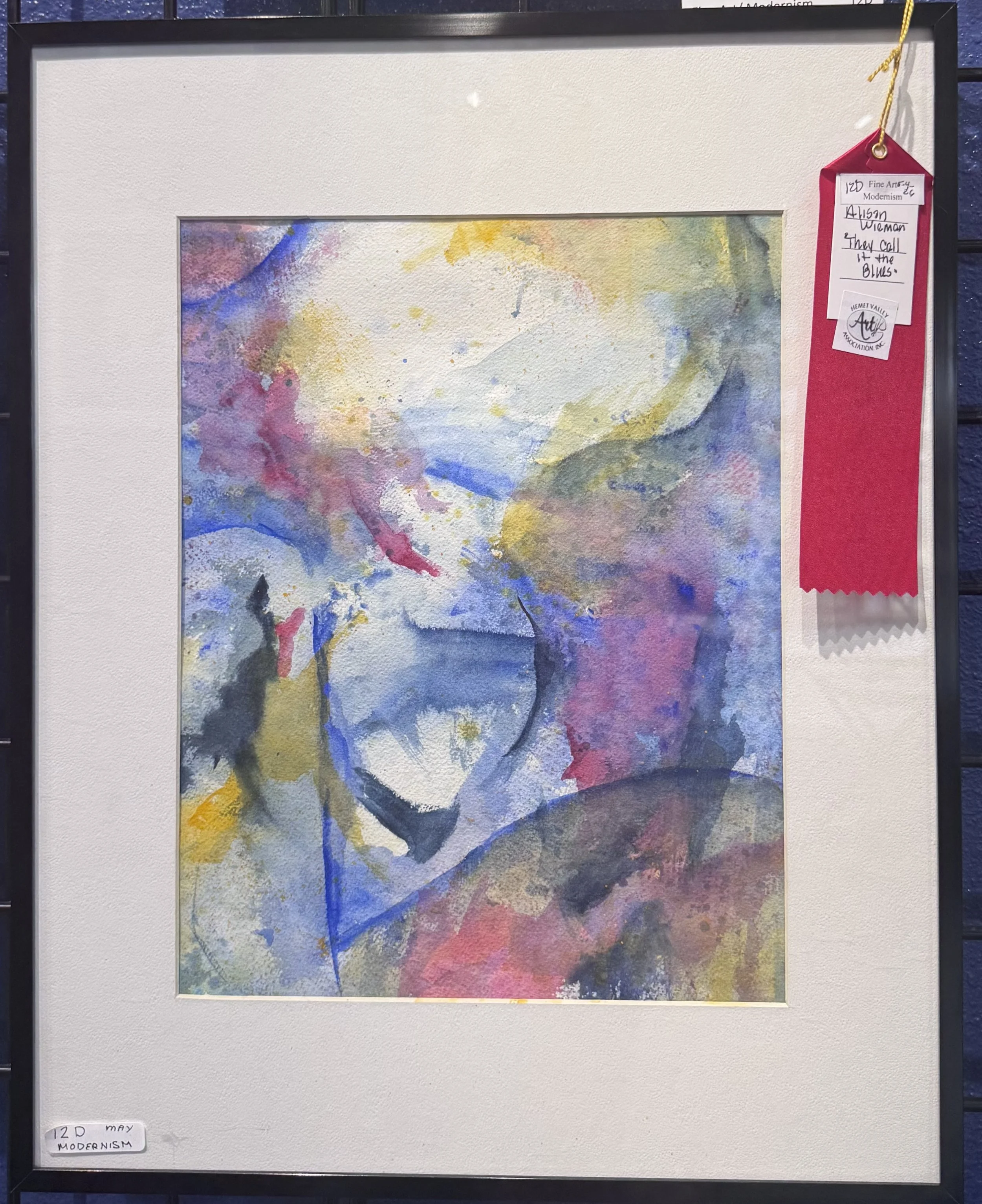 Watercolor painting with abstract swirls of yellow, blue, pink, and purple. Framed with a white mat and black border, with a red ribbon tag on the top right corner and a handwritten label in the bottom left corner reading '12D May Modernism.'