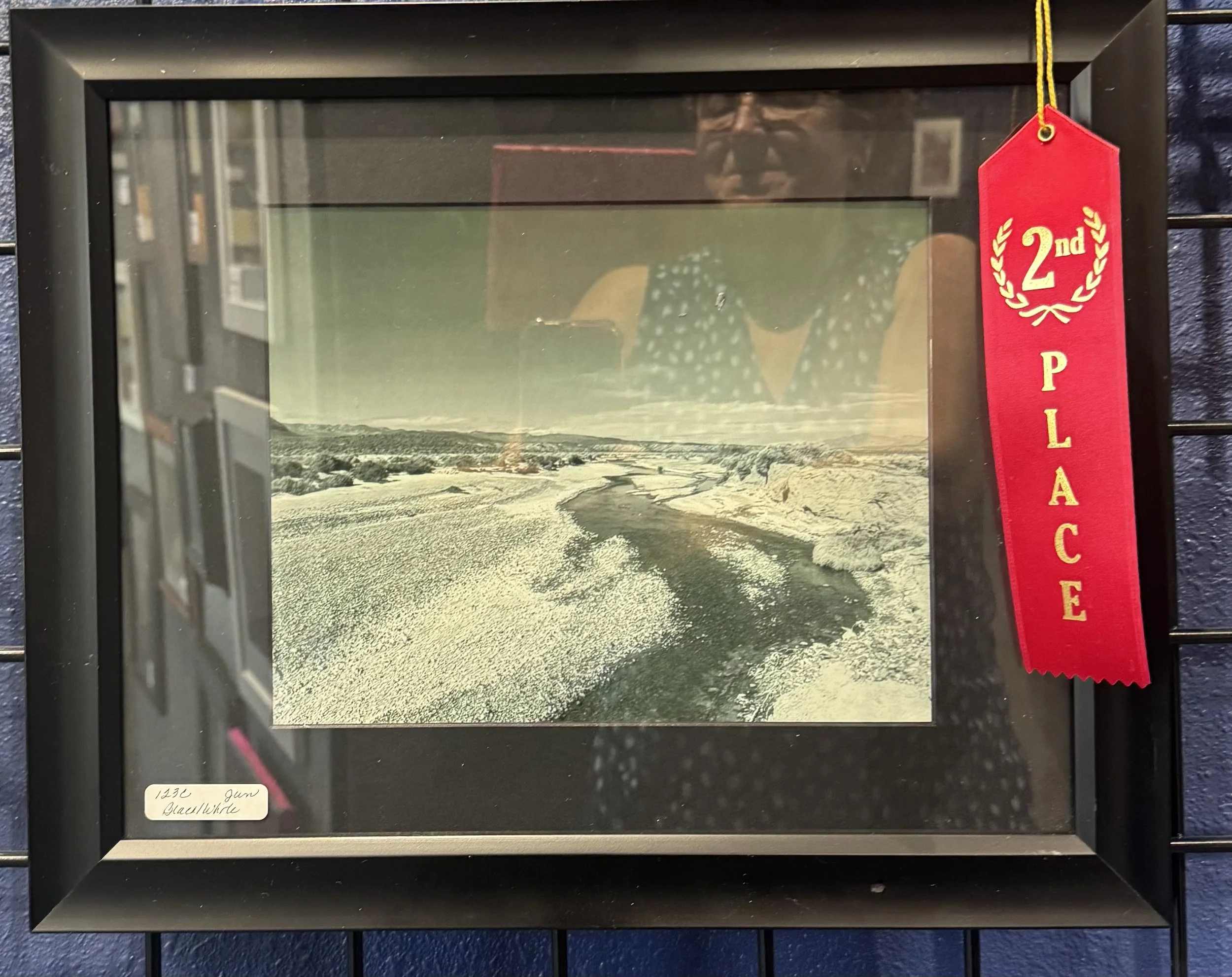A framed landscape photograph of a snow-covered river with ice patches and a bridge in the background, displayed inside a glass-fronted case with a red ribbon marked "2nd Place" hanging from the top right corner.