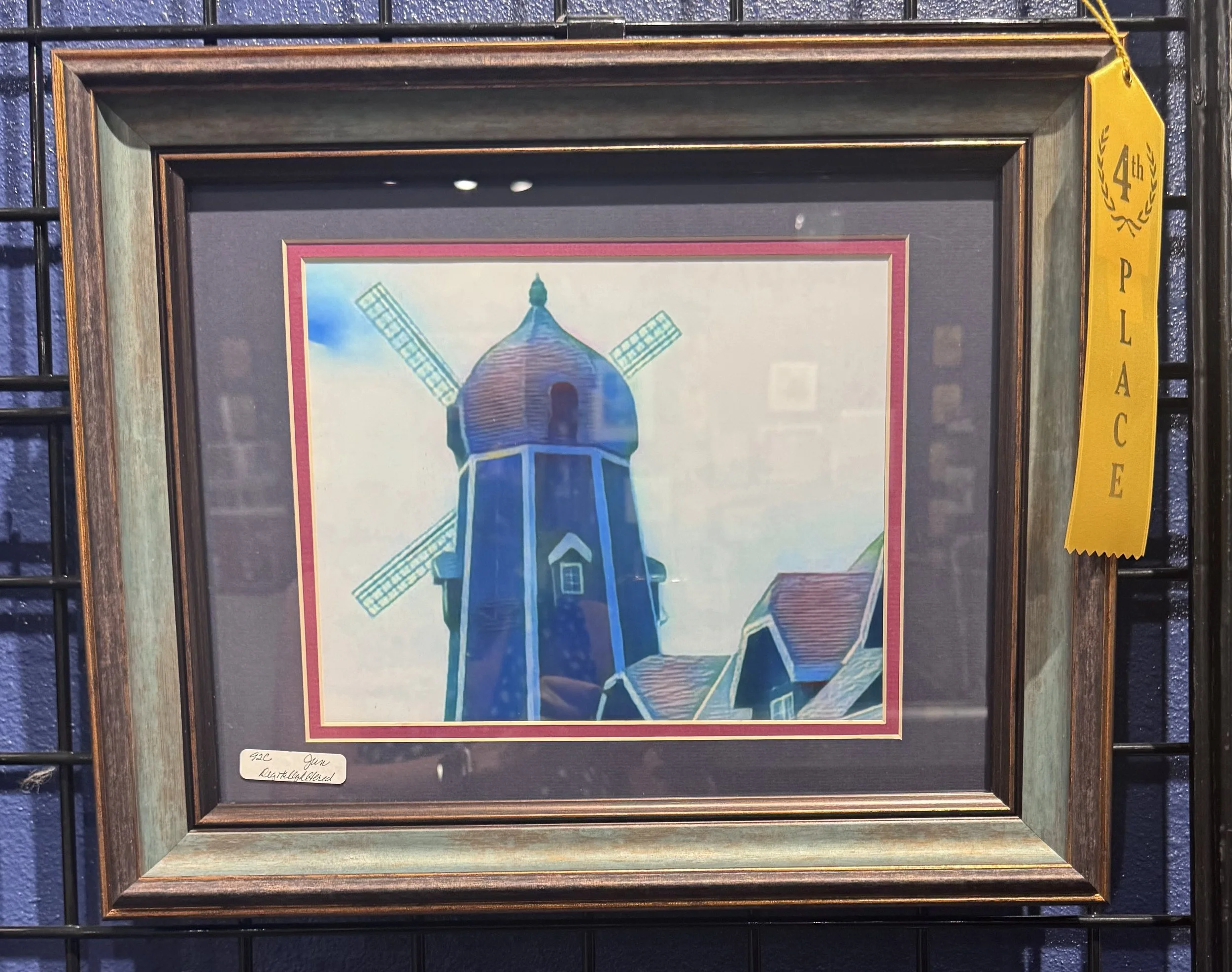 Framed artwork of a blue windmill with four blades and a rounded roof, set against a sky background with clouds, inside a wooden frame with a yellow exhibitor ribbon on the top right corner.