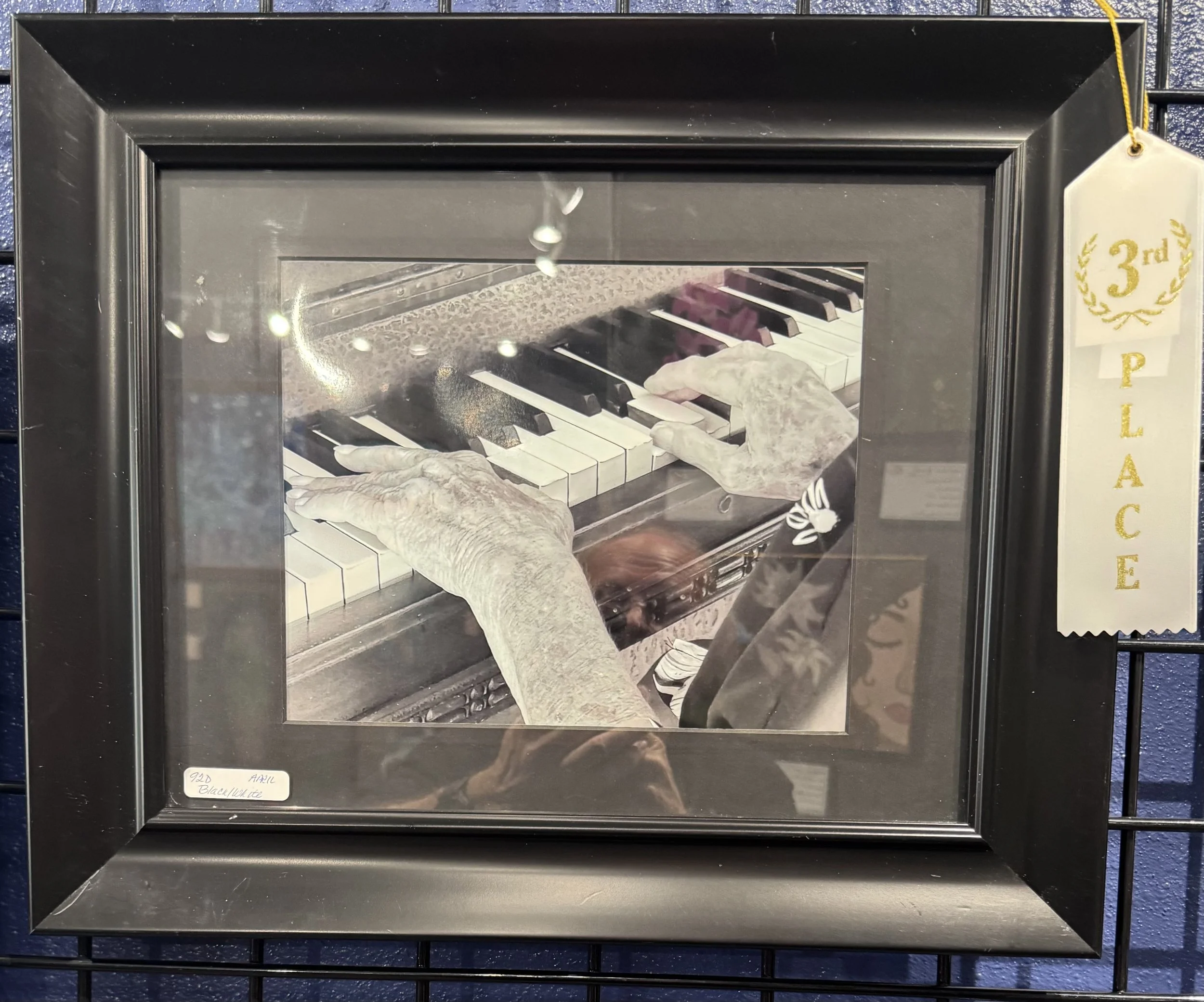 A framed black and white photograph of elderly hands playing the piano. The picture appears to be displayed at an exhibition, with a label underneath indicating the date as April 2020 and titled 'Beach Wite'.