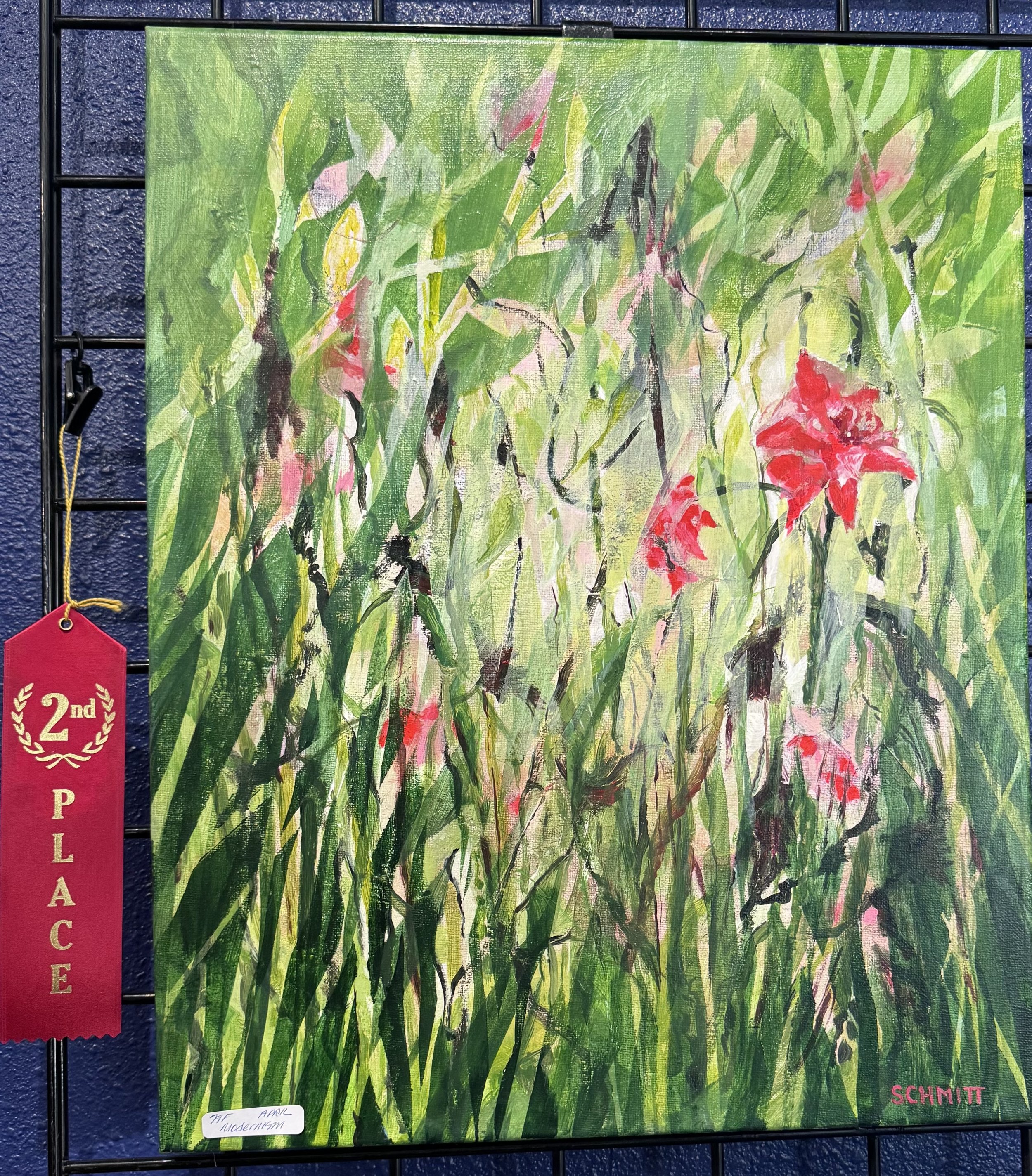 An abstract painting featuring green foliage with hints of pink and red flowers. The painting is displayed on a black grid wall with a red ribbon award indicating second place for the artist Schmitt, and a white label with handwritten text reading 'T