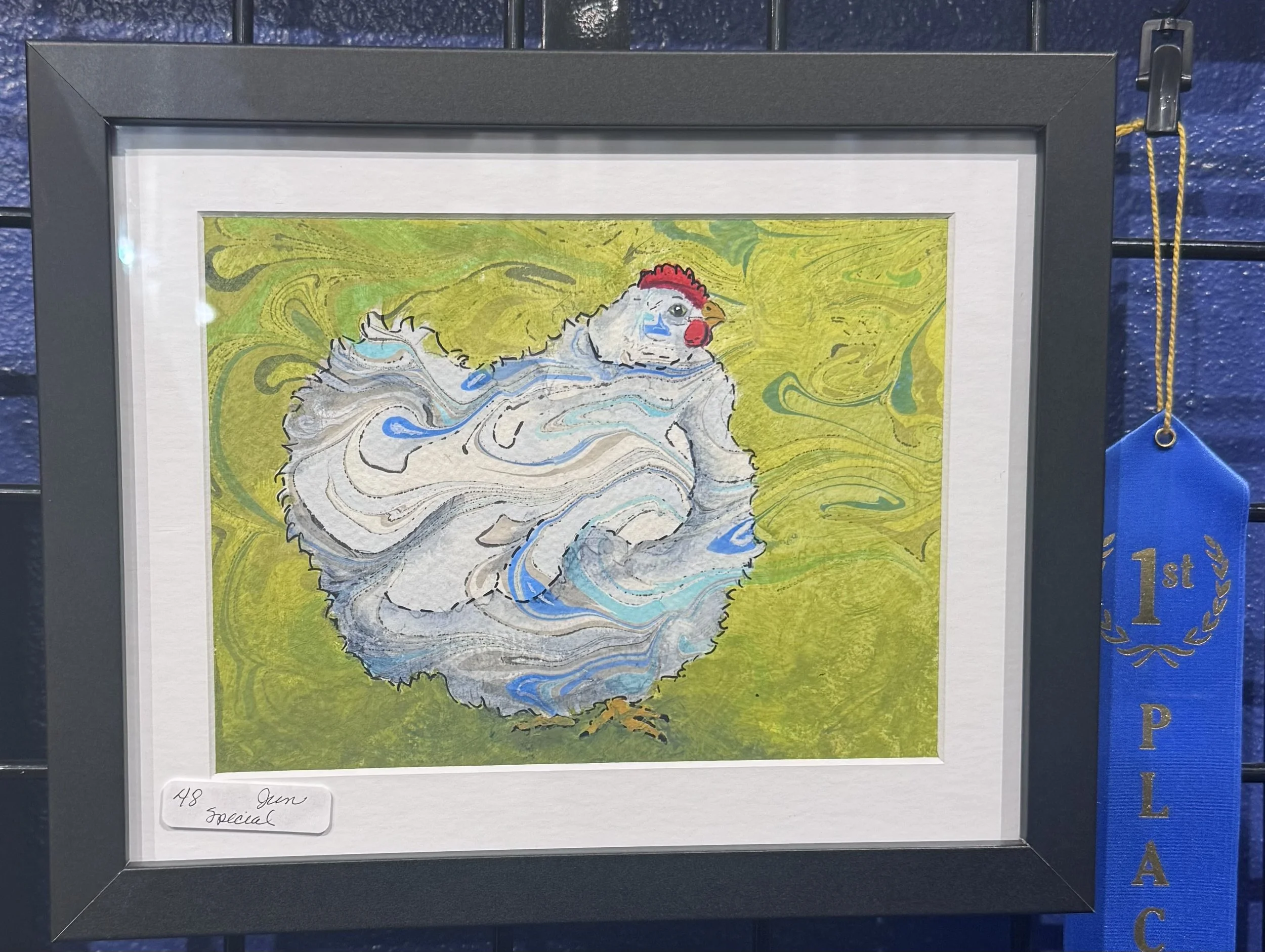A framed watercolor painting of a rooster with a vibrant red comb, gray and white feathers, standing against a yellow and green swirling background. The frame has a black border and a blue ribbon with a gold laurel design on the side indicates it is 