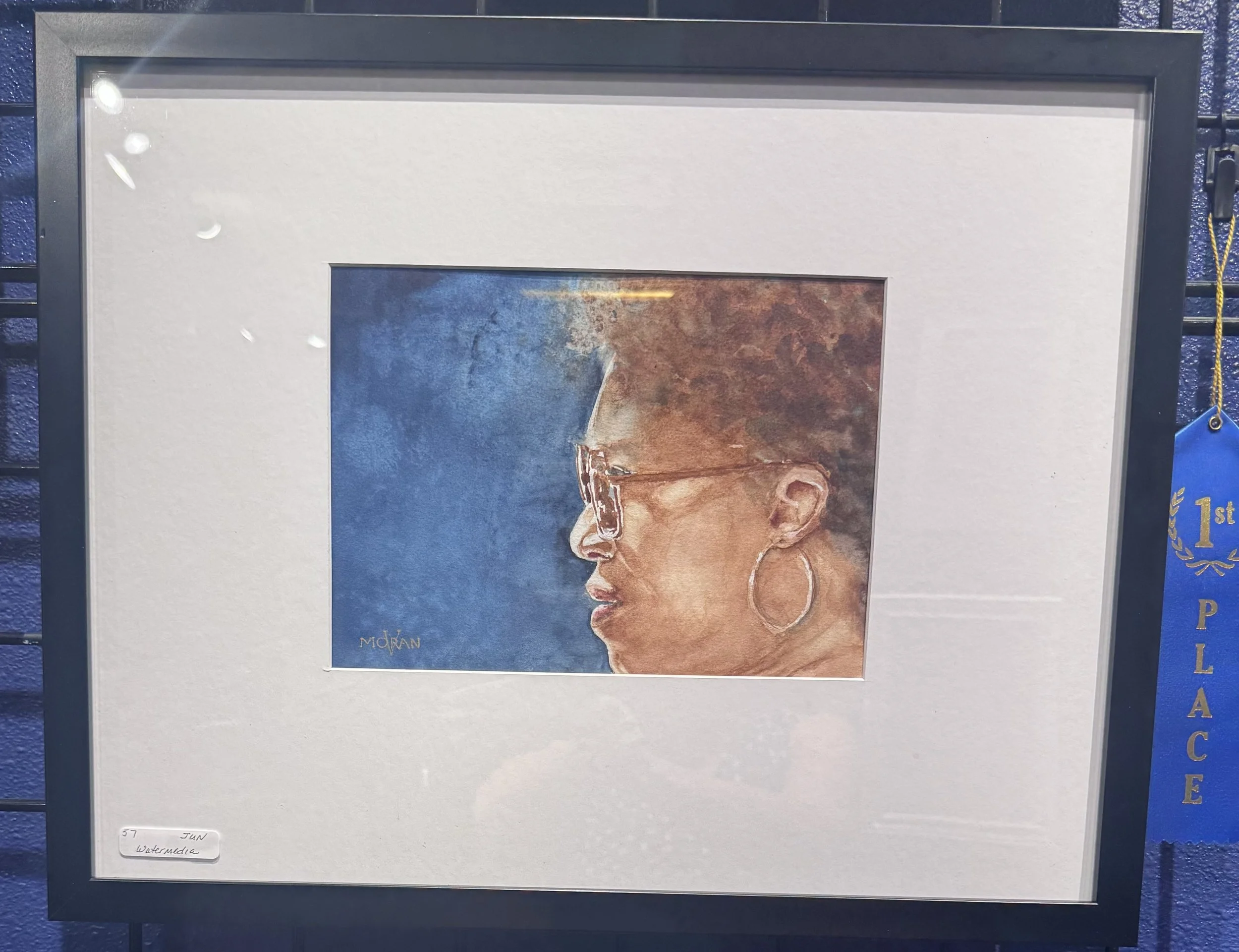 Close-up of watercolor painting featuring a side profile of a woman with glasses and large hoop earrings, with textured brown hair and a blue background.