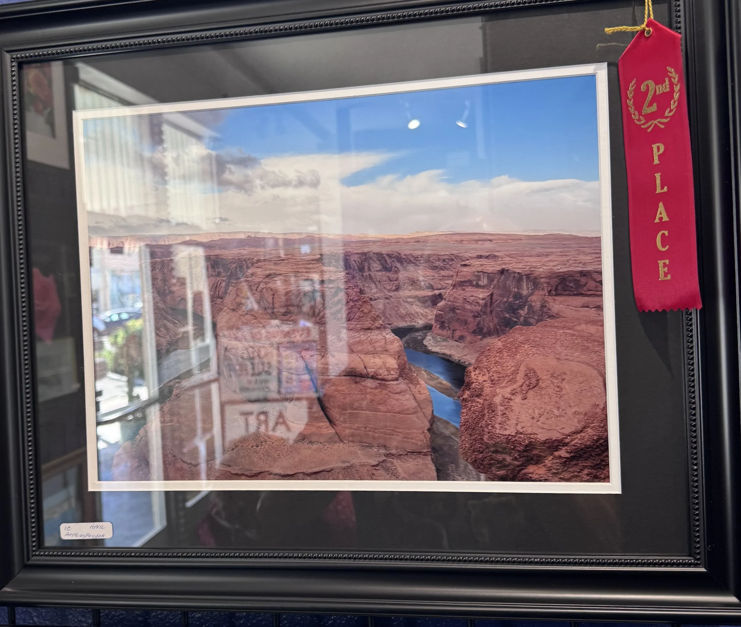 Photograph of a framed landscape image of the Grand Canyon with red rock formations and a blue sky with clouds, displayed inside a black frame with a pink ribbon indicating second place.