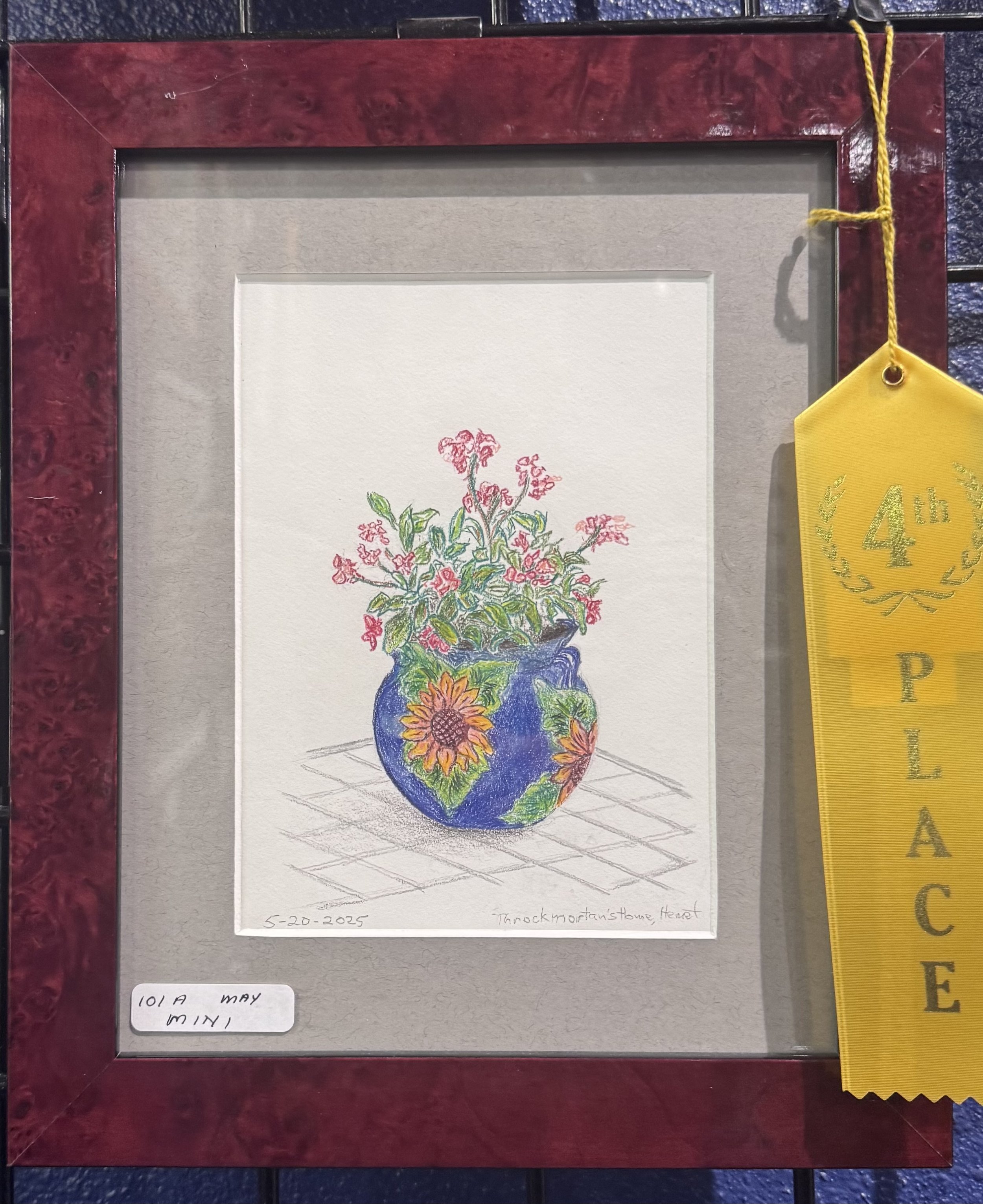 Colorful drawing of a potted plant with pink flowers, green leaves, and yellow flowers at the bottom, on a white piece of paper with handwritten text and date. The drawing is framed and displayed on a surface, with a yellow ribbon on the right side.