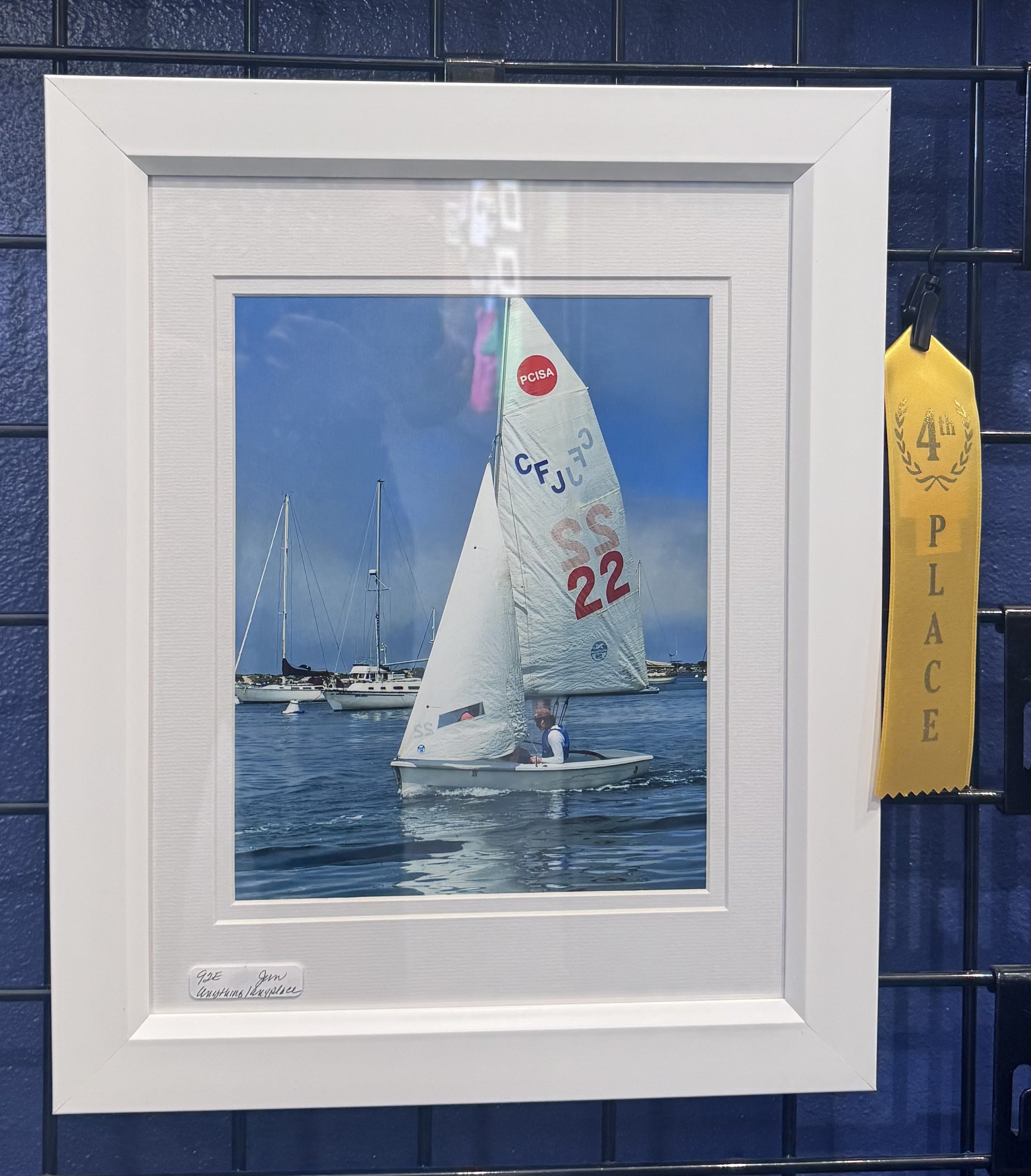 A framed photograph of a sailboat on calm water, with blue sky and other sailboats in the background. The sailboat has a white sail with the number "22" and the letters "CFJC" visible. To the right of the frame, there is a yellow ribbon with the text