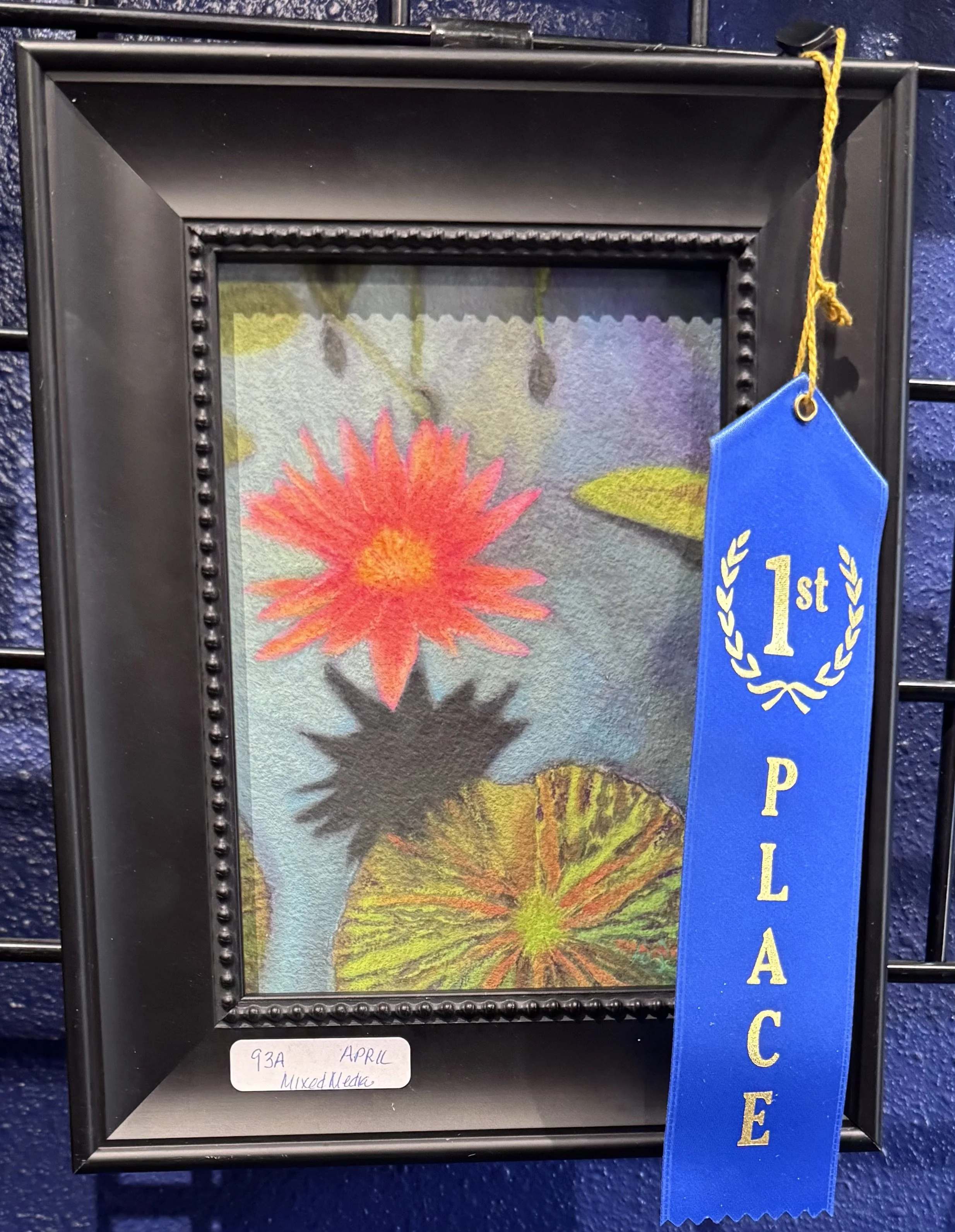 A framed watercolor painting of colorful flowers, mostly in shades of pink, orange, yellow, and green, displayed on a blue textured wall with a blue ribbon labeled '1st Place' hanging on the right side.