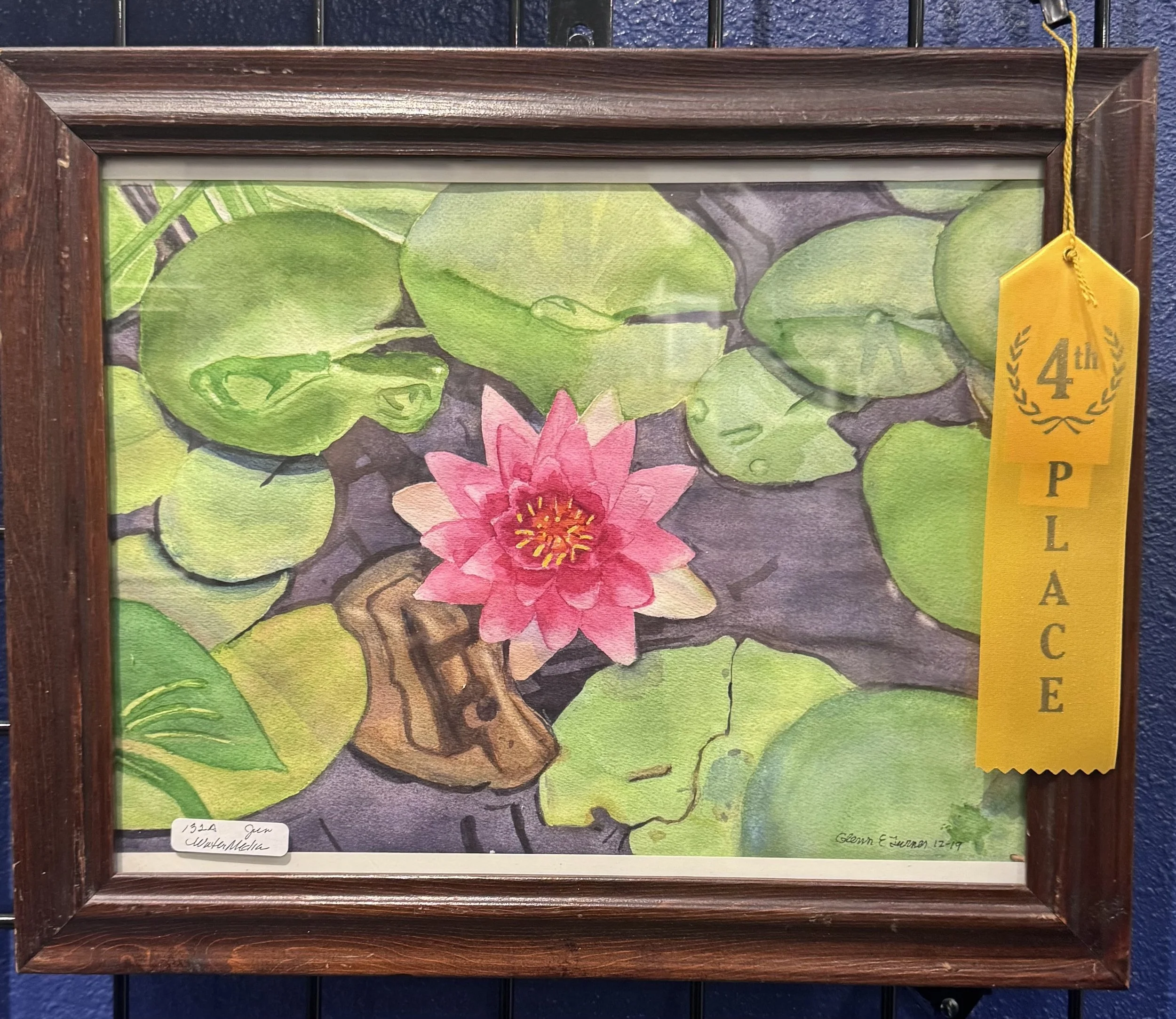 Framed artwork of a green frog on lily pads with a pink water lily flower, having a yellow ribbon award tag on the top right corner.