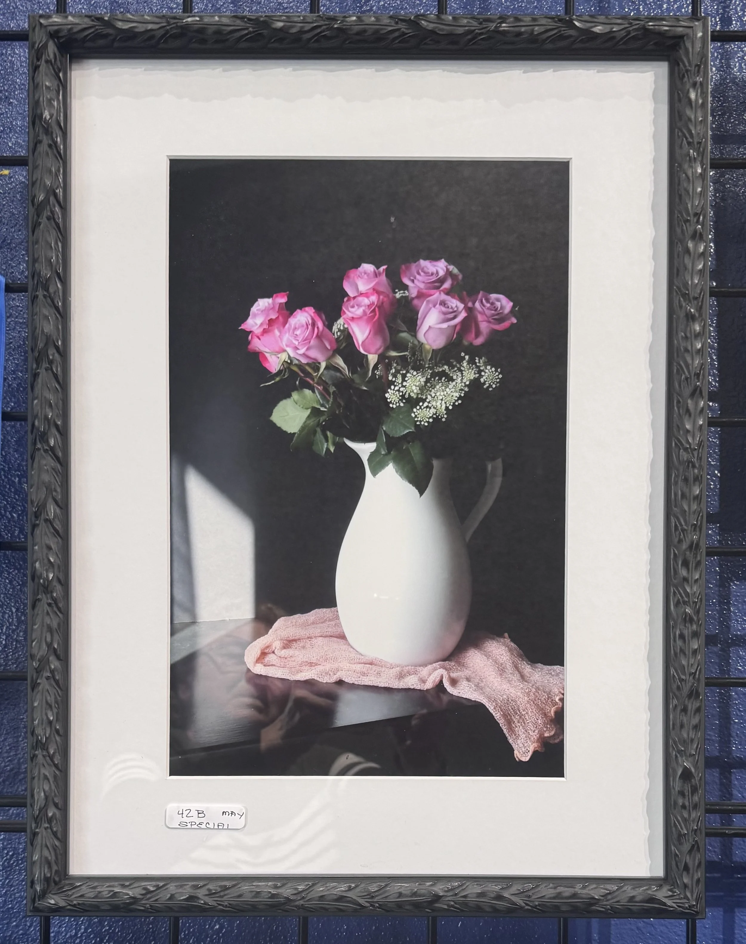A framed photograph of a bouquet of pink roses in a white vase with baby's breath flowers, placed on a pink lace cloth, against a dark background.