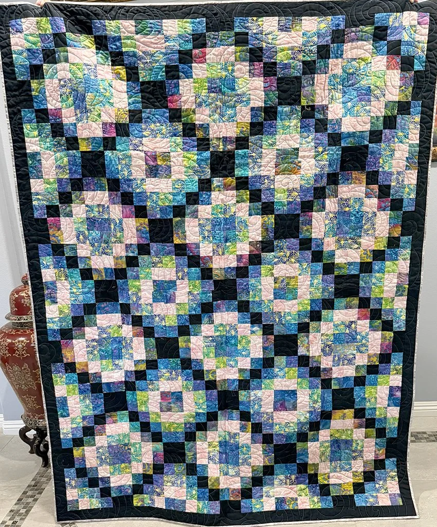 A colorful quilt with a pixelated floral pattern in pastel and bright colors, framed with a dark border.
